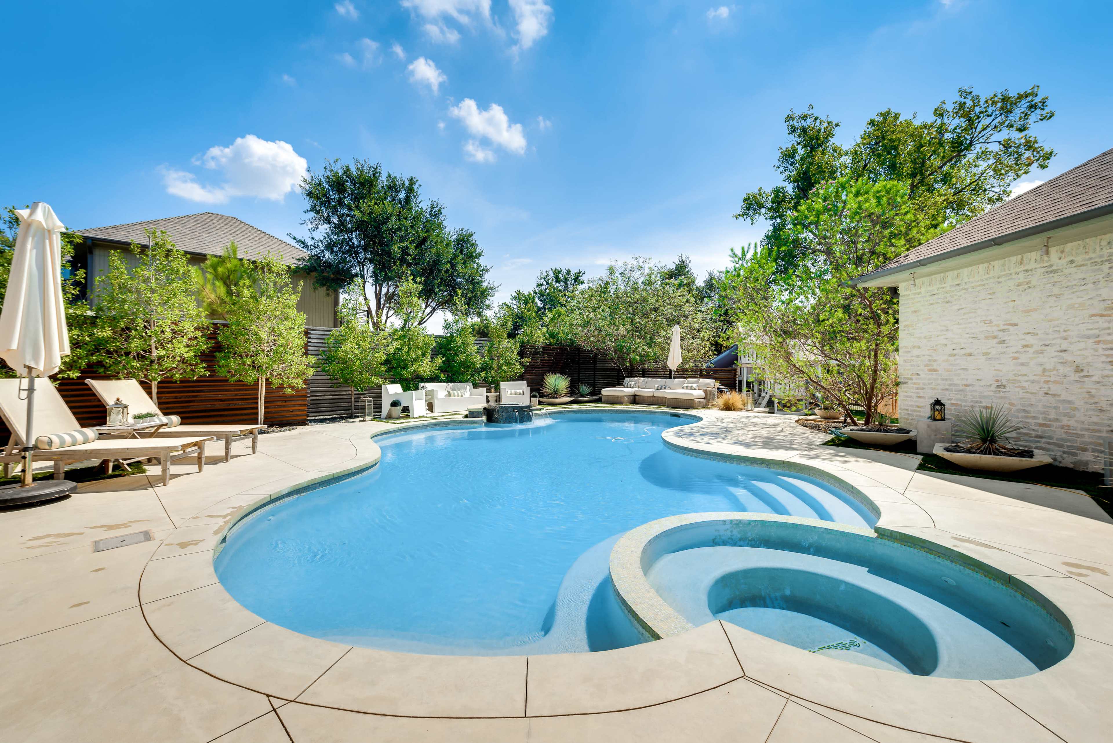 The image shows a residential backyard featuring a large, shaped swimming pool with a spa area, surrounded by greenery and patio furniture.
