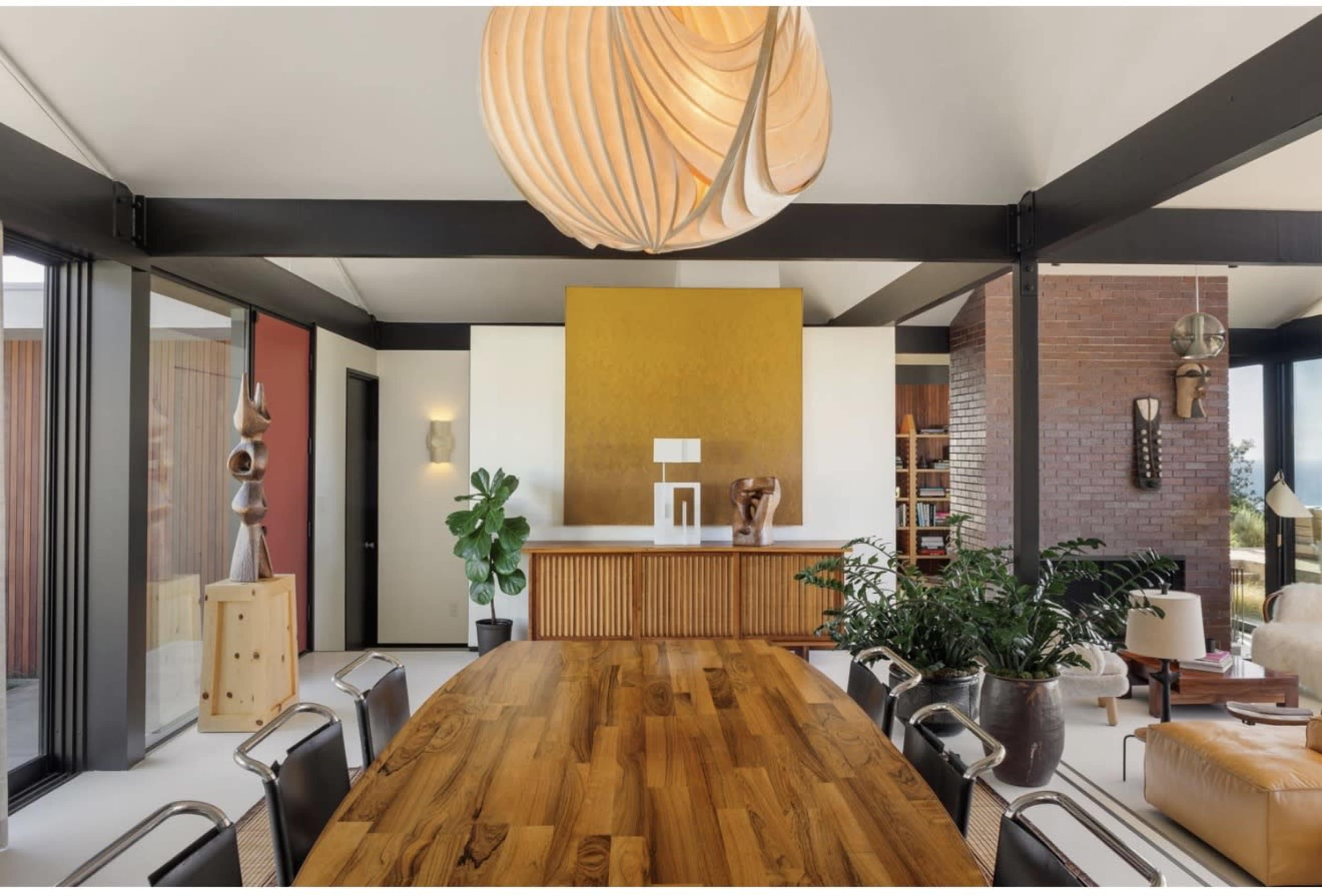 The image shows a modern dining area featuring a large wooden table, a sculptural light fixture, and a backdrop of shelving and decorative elements.
