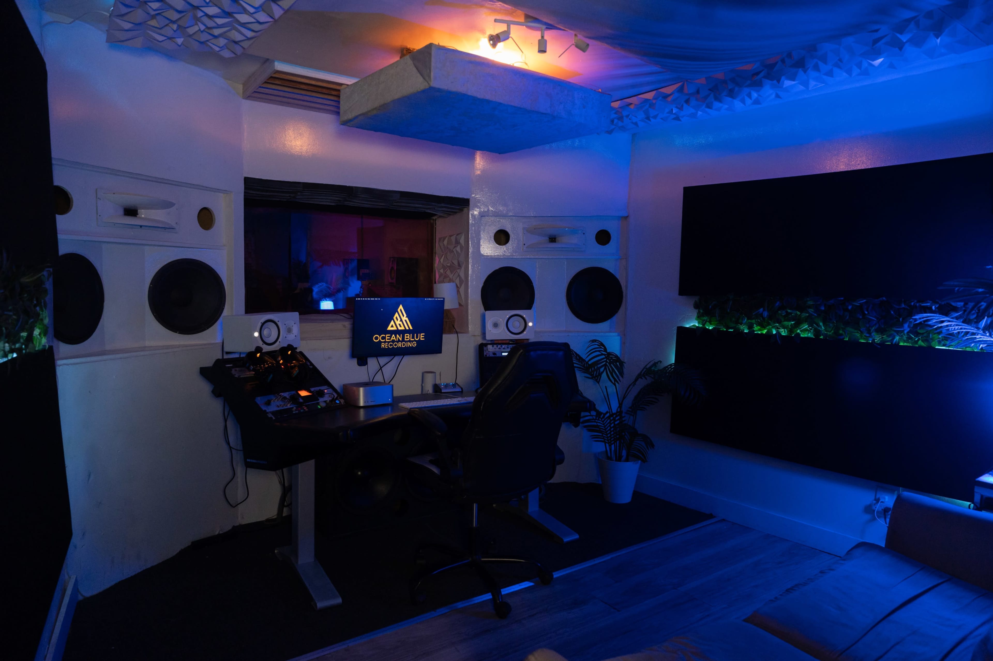 The image depicts a music studio featuring a large desk with audio equipment, speakers mounted on the walls, and ambient blue lighting.