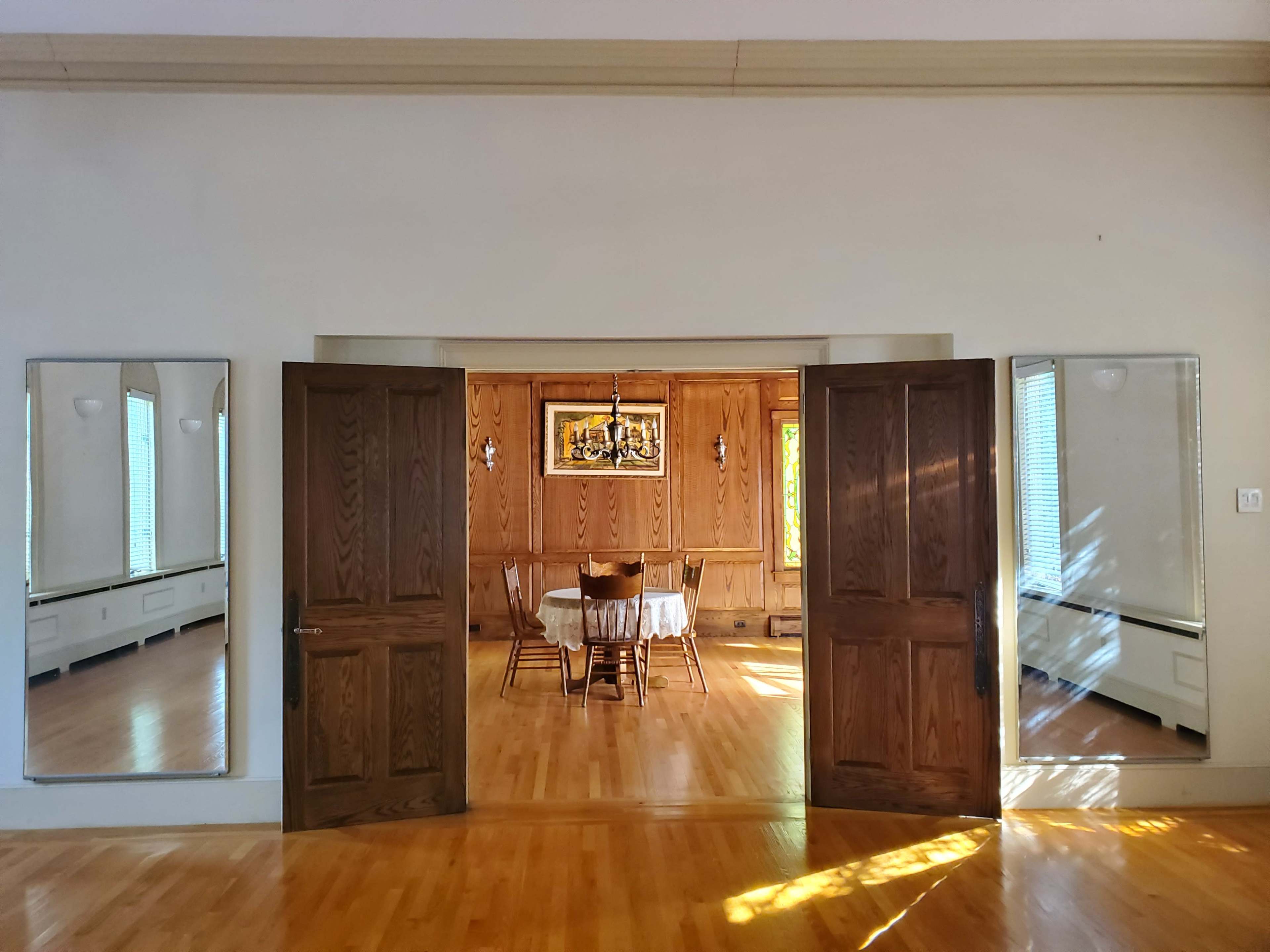 A spacious room with large wooden double doors leading to a warmly-lit dining area furnished with a table and chairs, framed by two mirrors on either side.