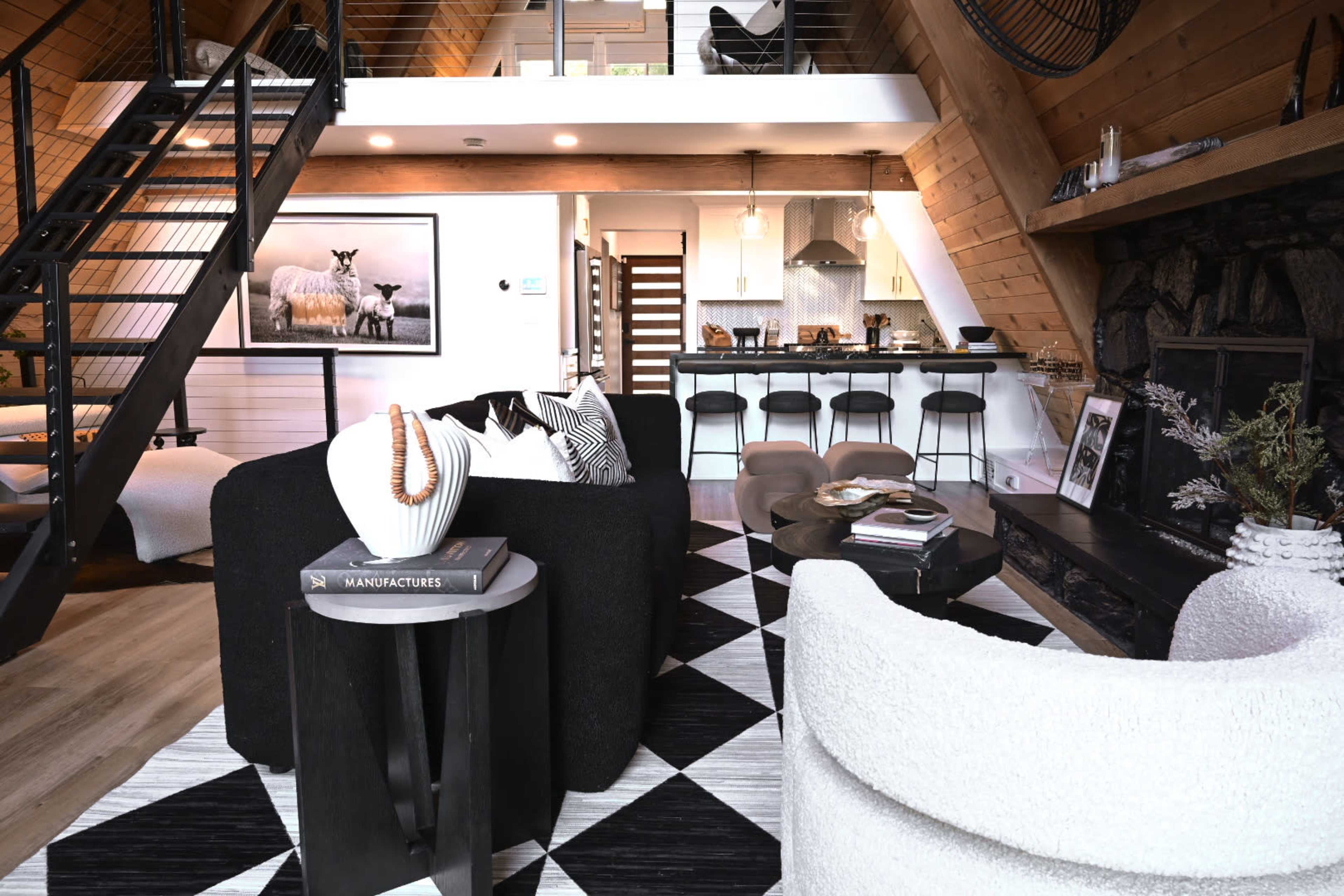 A modern living room with a staircase leading to an upper level, a large fireplace, and a kitchen area visible in the background, all decorated in a stylish black and white color scheme.