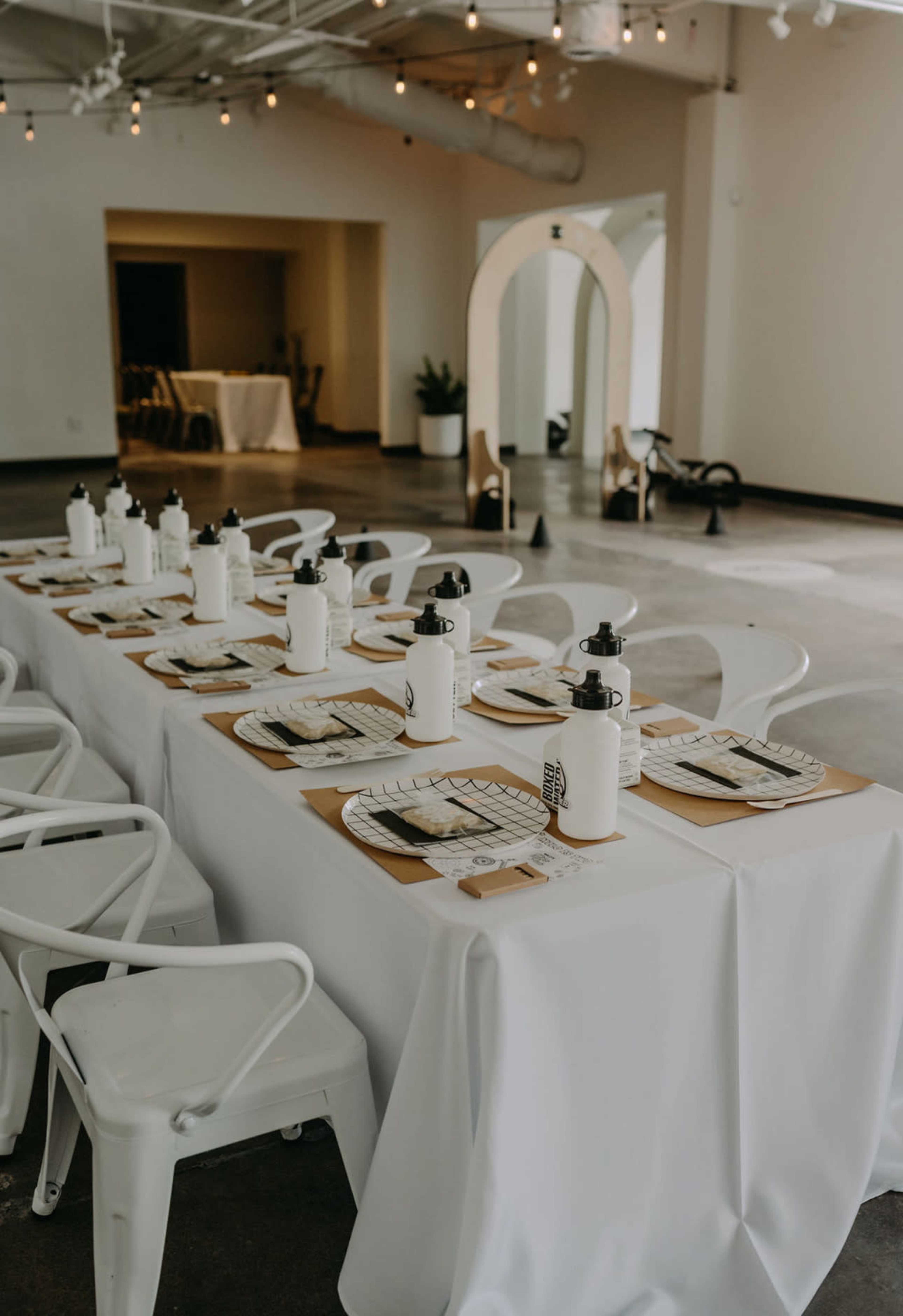 A long dining table is set with white tablecloths, plates, and bottles, arranged in a well-lit, minimalist interior space.