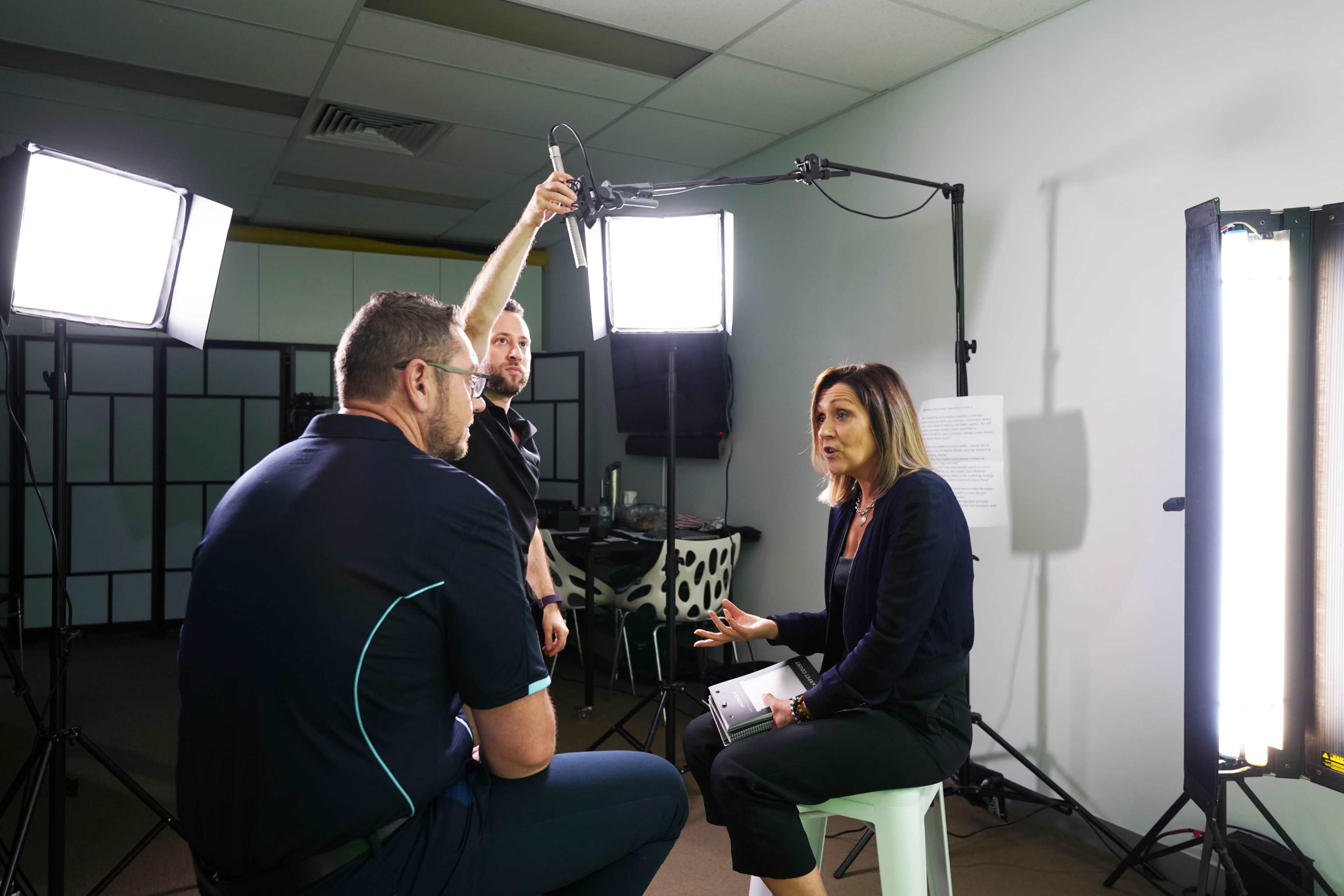 A woman interviews a man in a studio setting with three bright lights and a microphone overhead.
