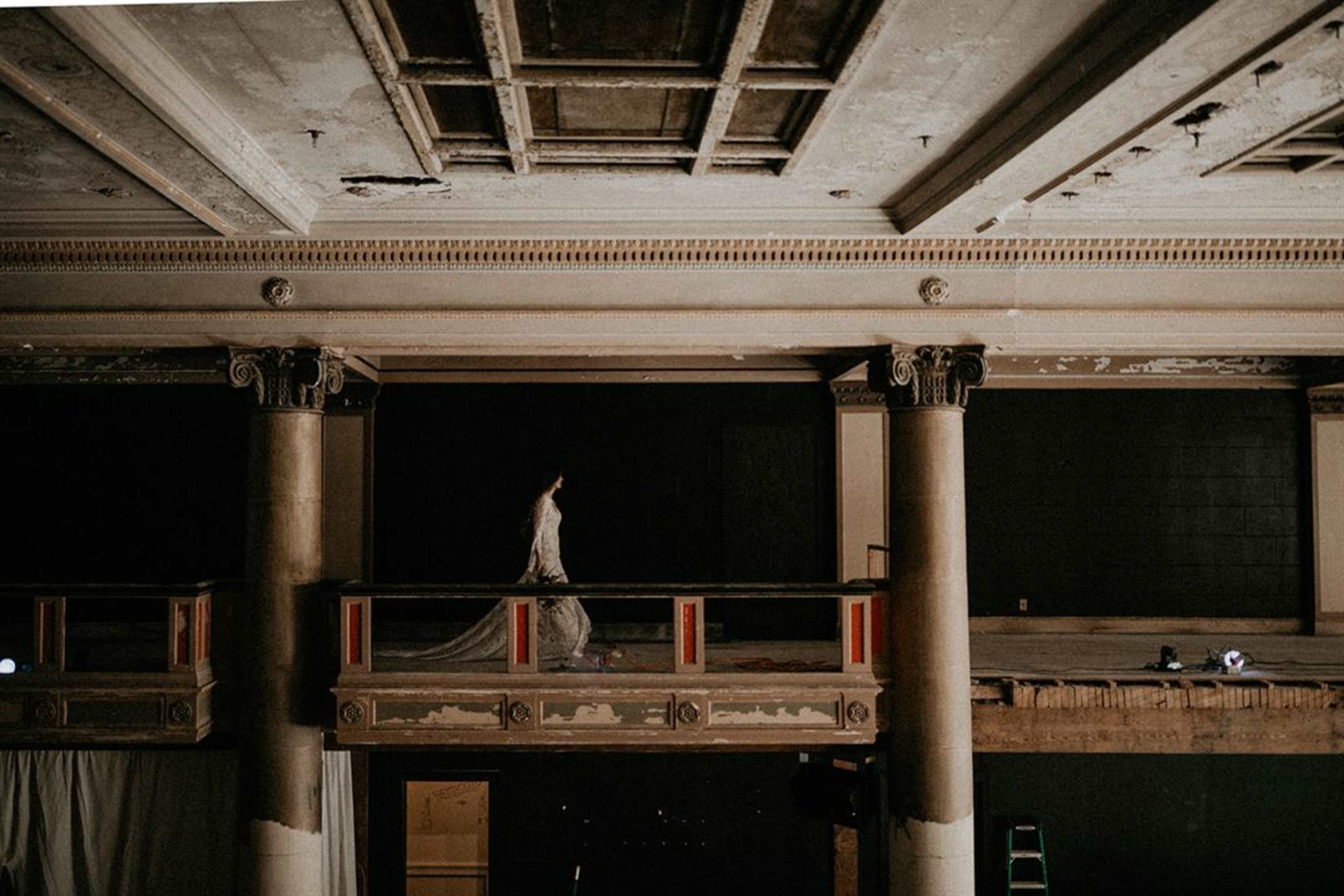 A figure walks along the balcony of an elegant, historic building with ornate columns and a decorative ceiling.