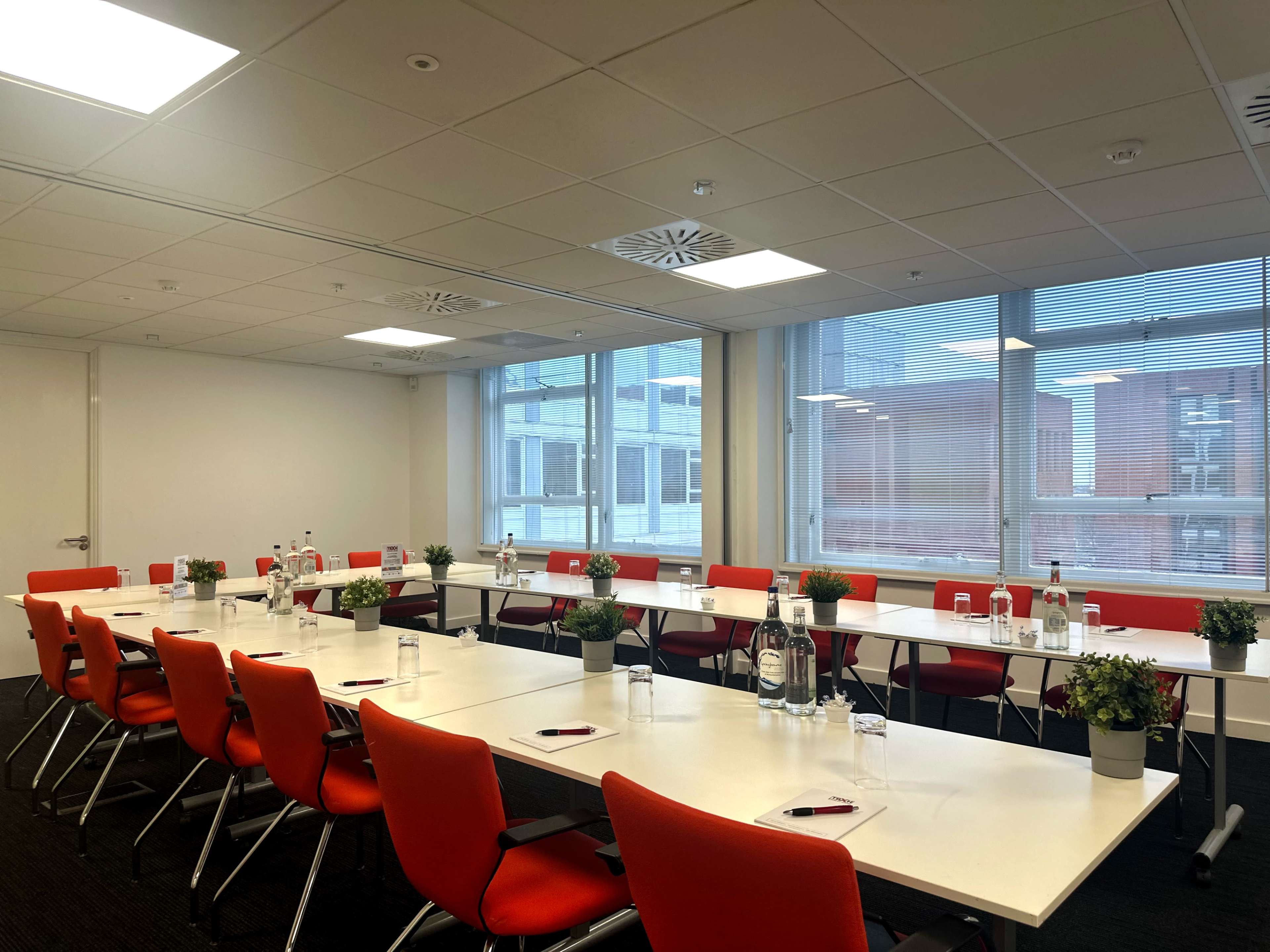 A long conference table is set up with red chairs, water bottles, and small plants, in a well-lit meeting room featuring large windows.