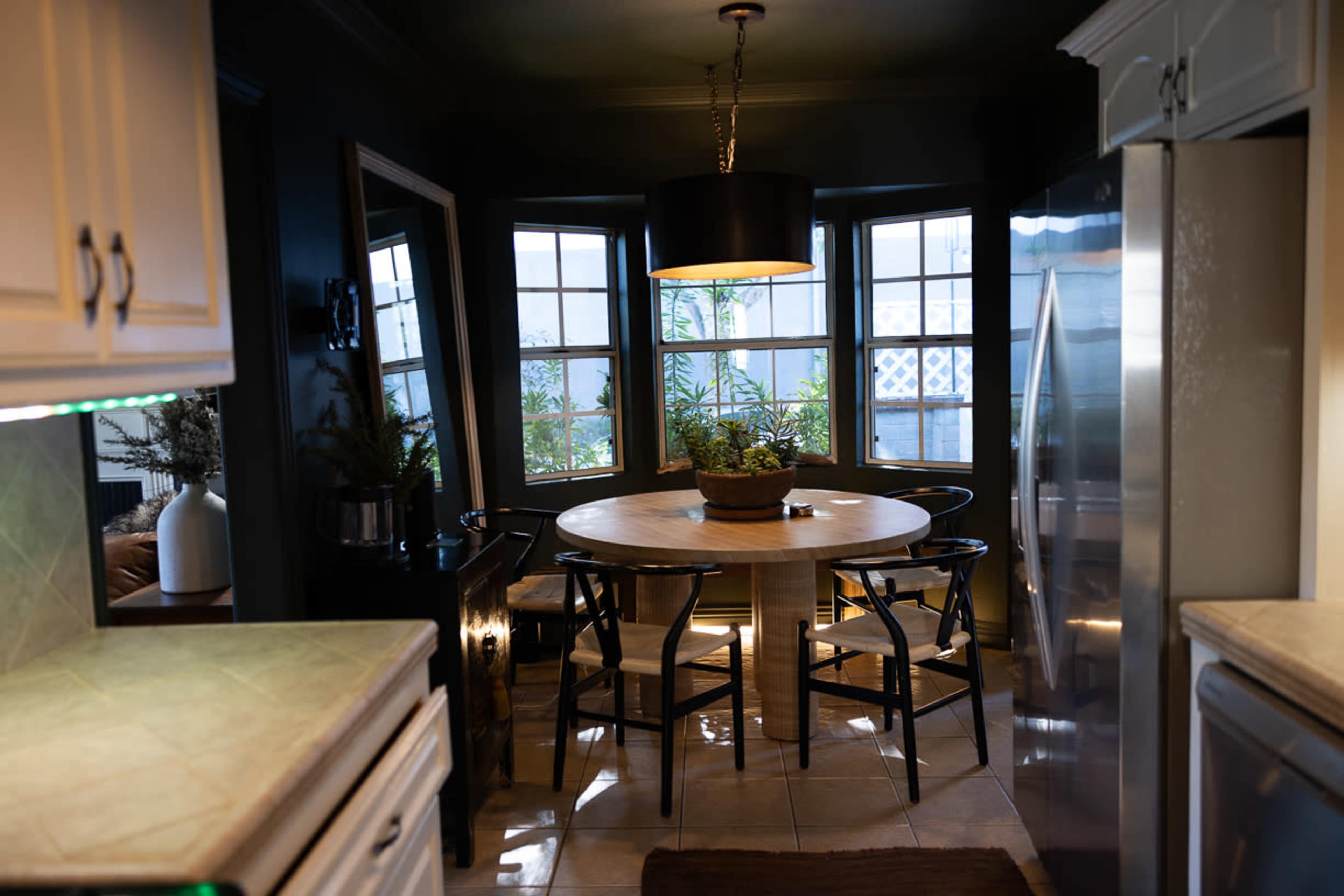 The image shows a modern kitchen with a round wooden table and four chairs, surrounded by large windows allowing natural light to enter.
