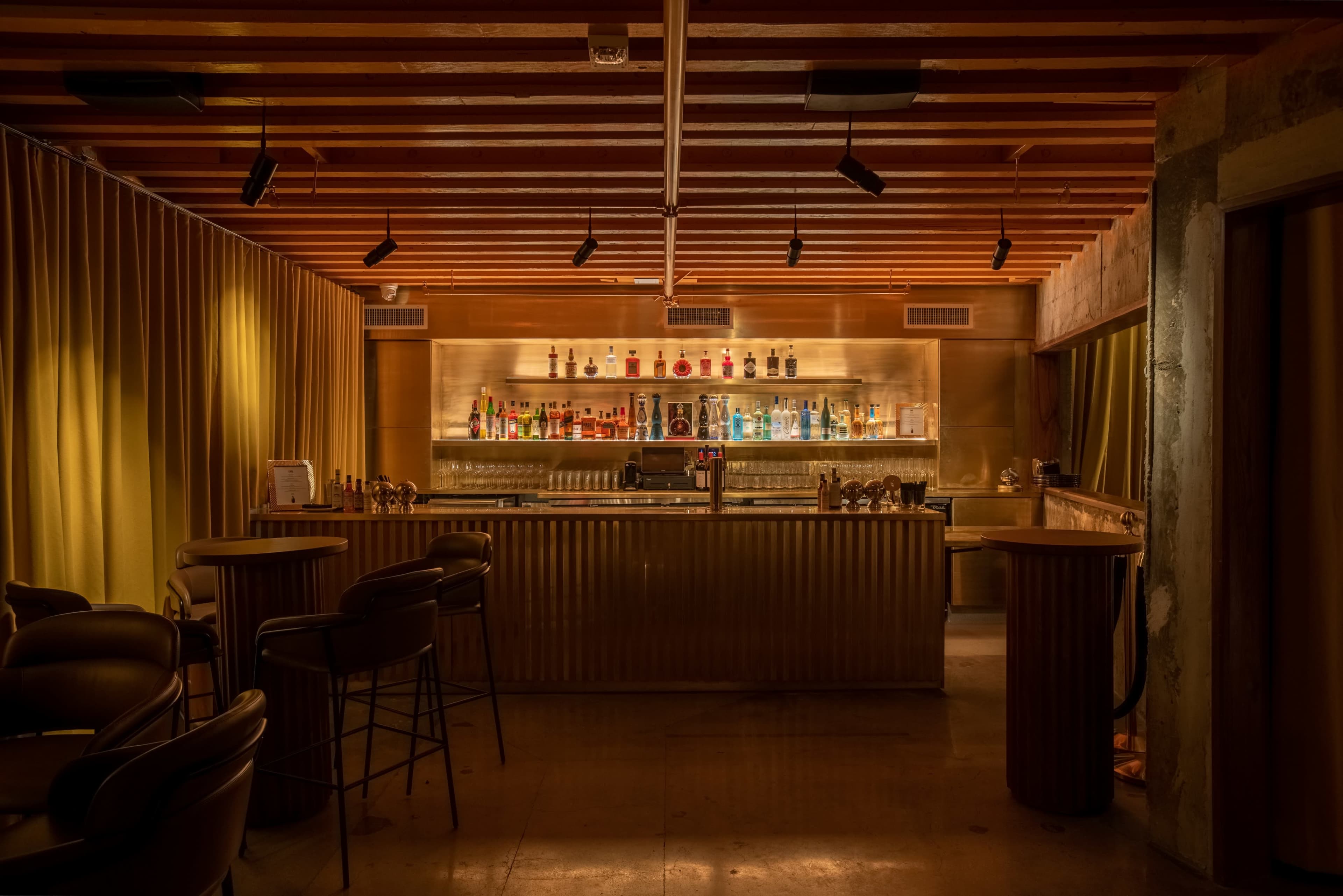 The image shows a dimly lit bar with a polished counter, shelves lined with colorful bottles, and a cozy seating area featuring black bar stools.