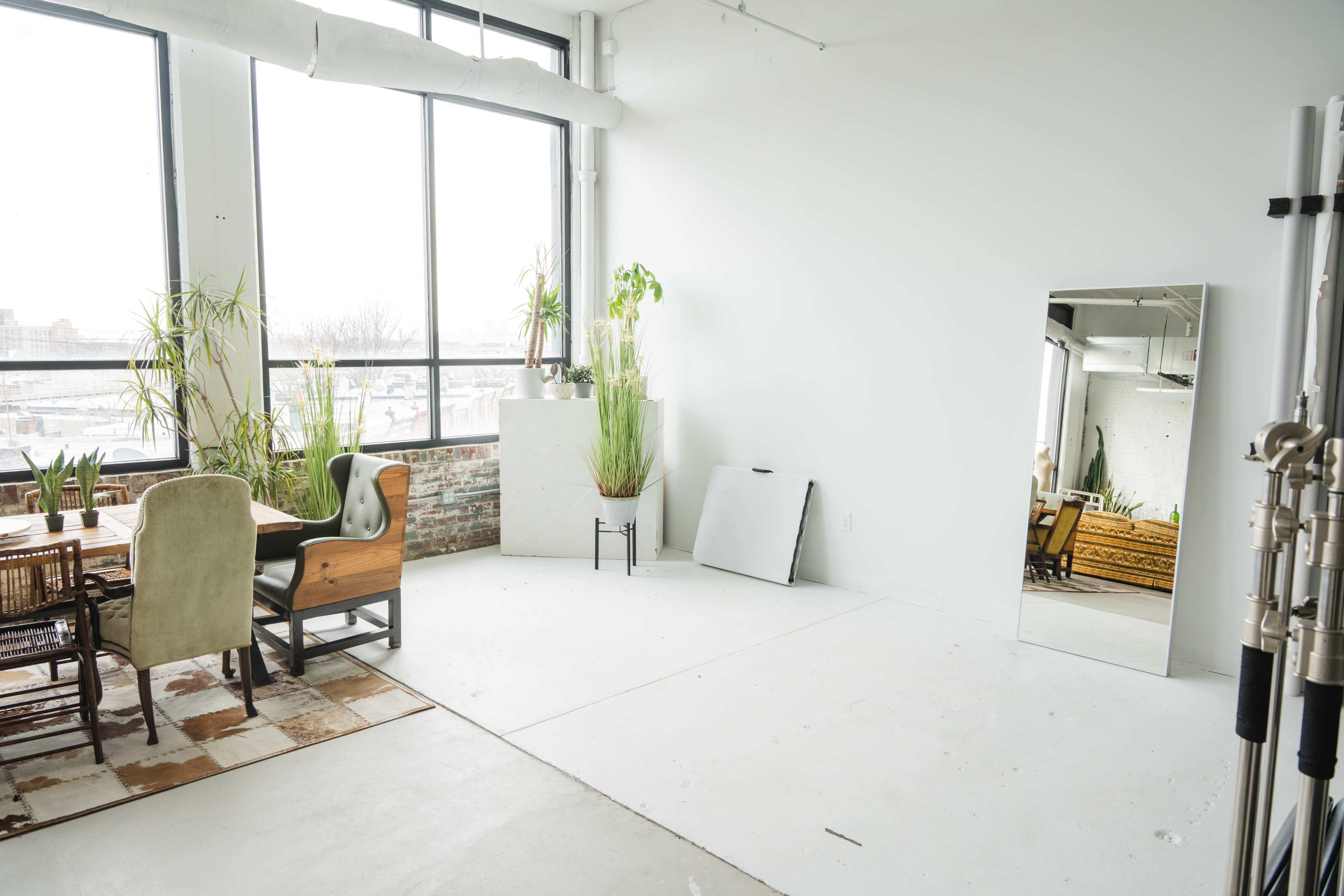 A spacious, minimalist interior with large windows, a wooden dining table, chairs, and several plants arranged around a mirror and a clear floor area.