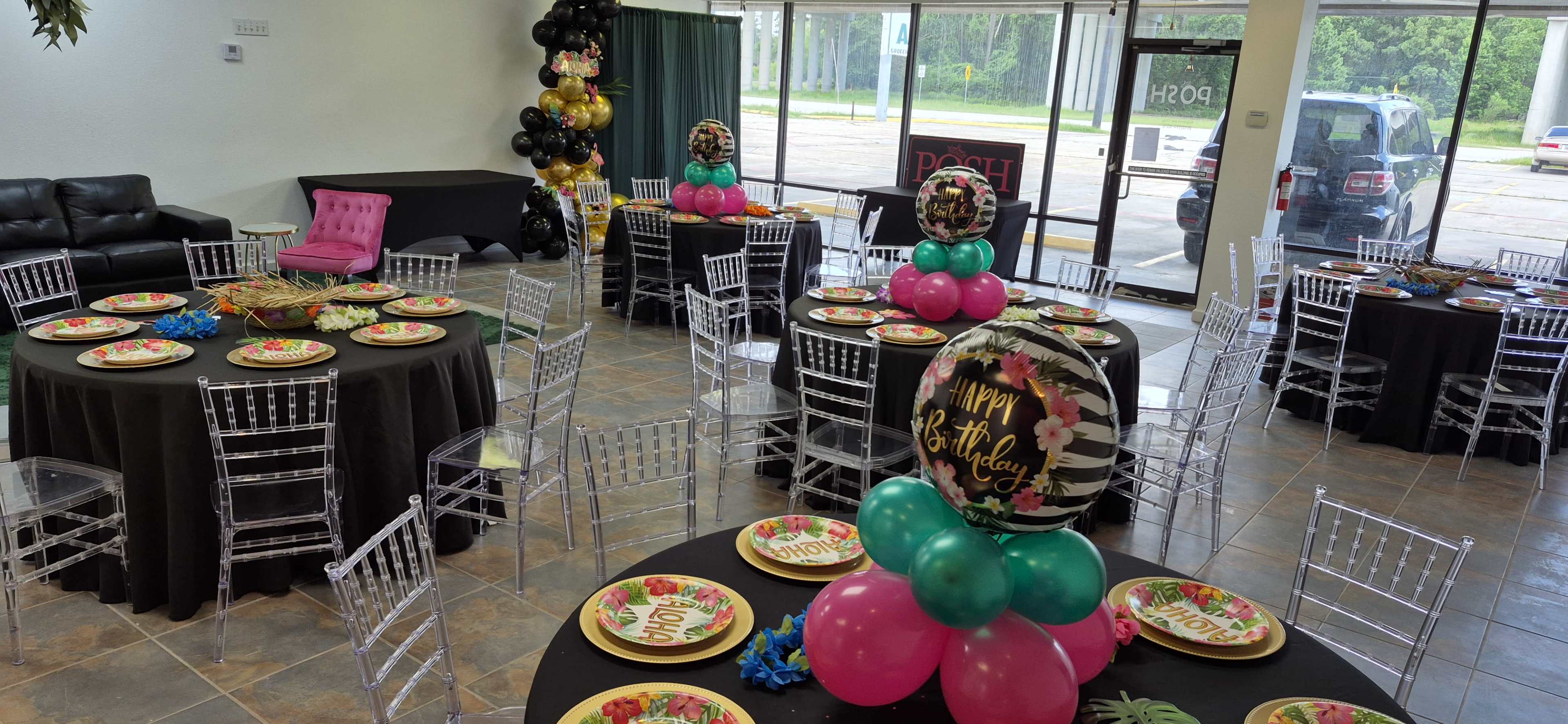 The image shows a decorated event space with round tables covered in black cloth, each set with colorful plates and adorned with balloon centerpieces, ready for a birthday celebration.