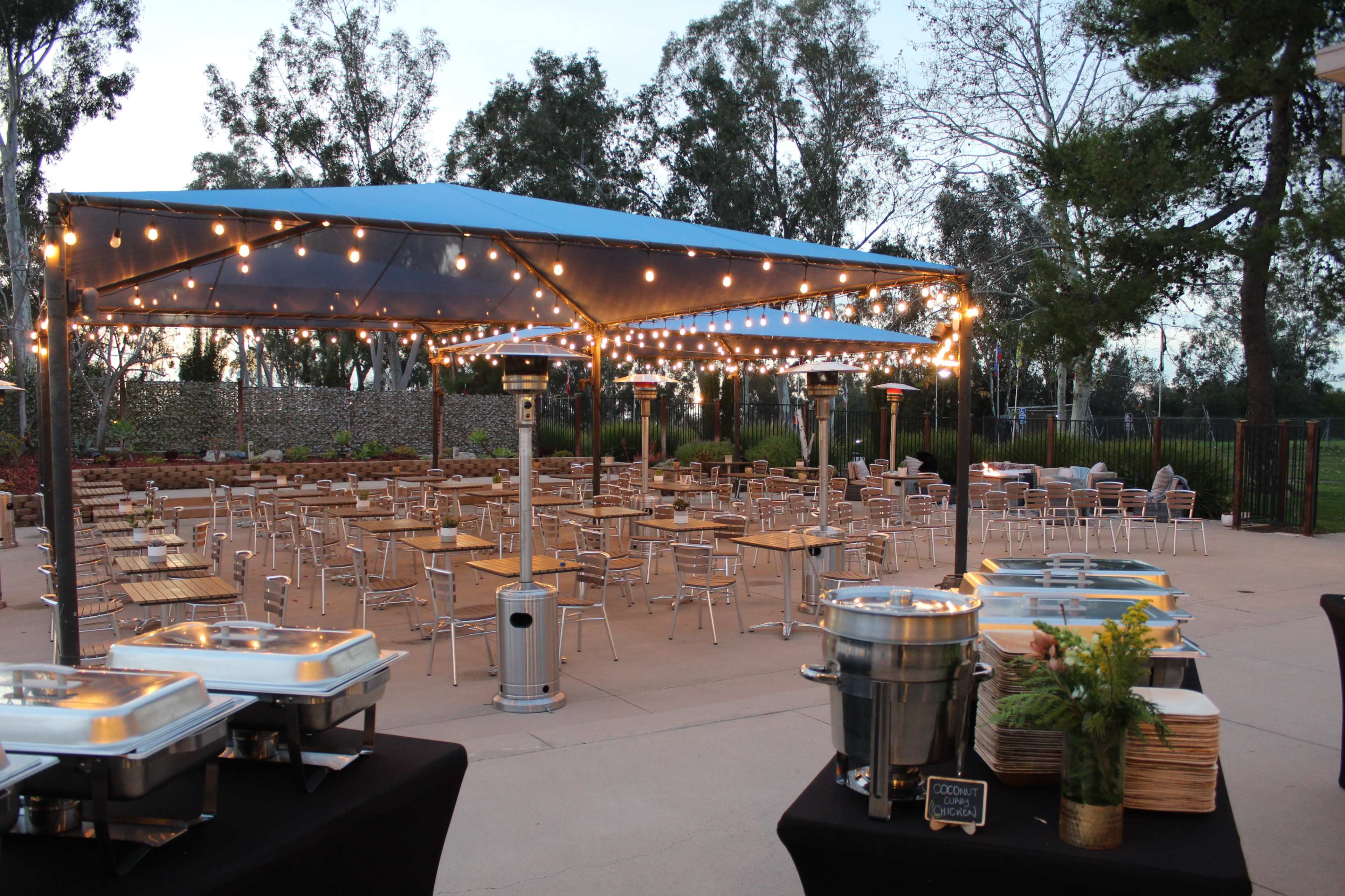 An outdoor dining area features a tented space with string lights, tables, and metal chairs arranged around buffet stations.
