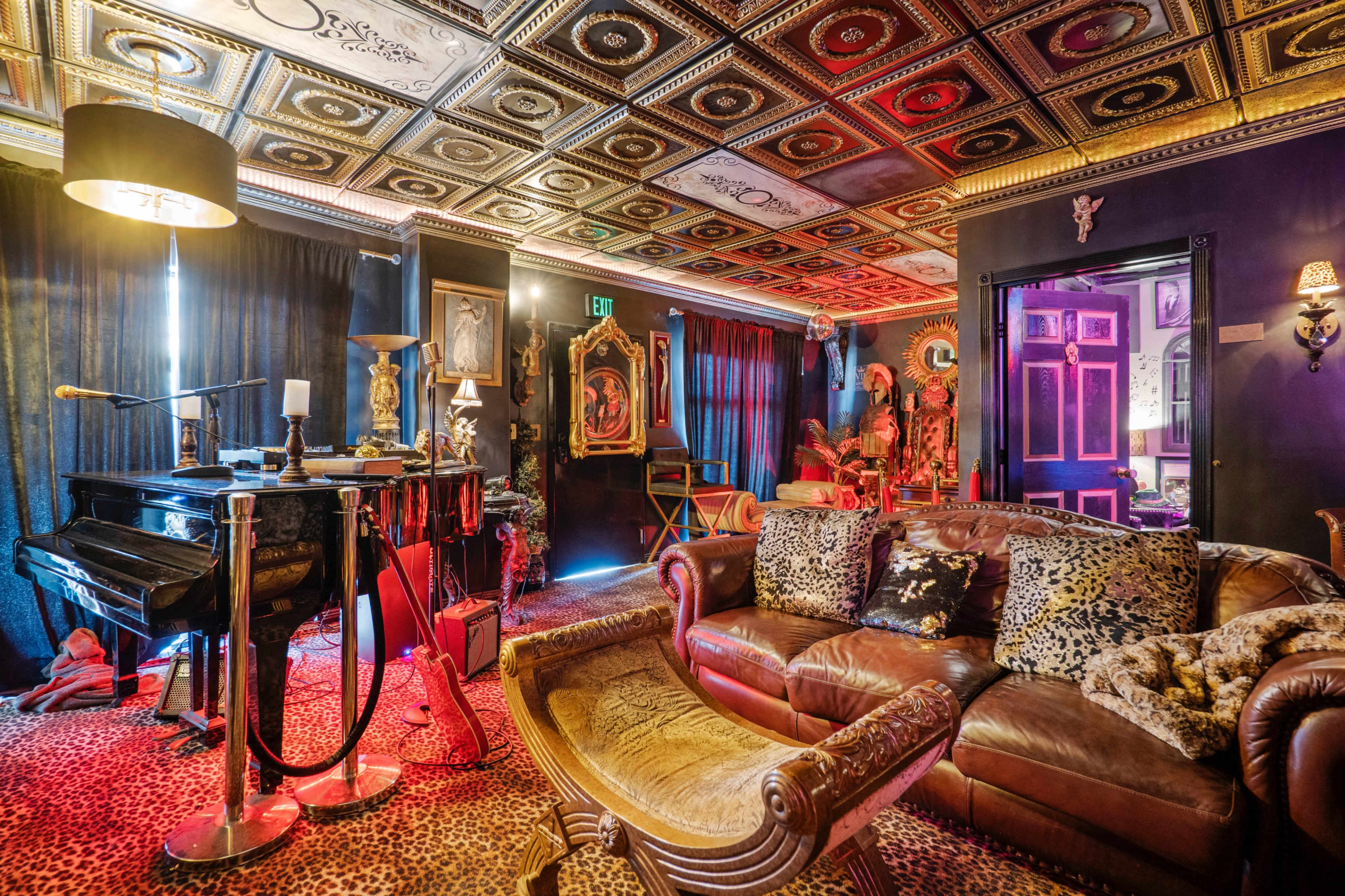 A richly decorated room featuring a piano, leather sofa, and ornate ceiling, illuminated by colorful lighting and adorned with various art pieces and furnishings.