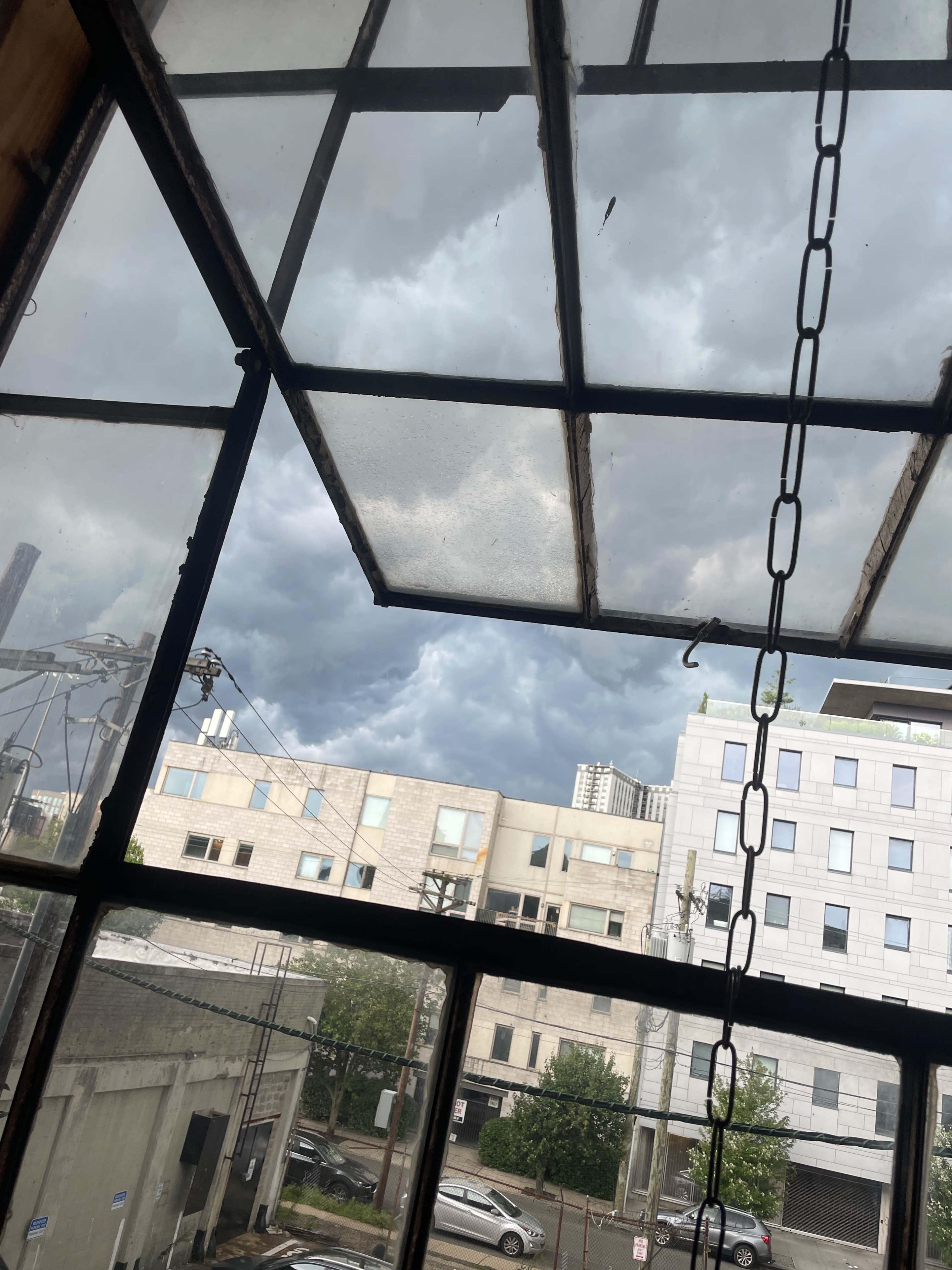 The image shows a view of dark, cloud-covered skies through a large window with metal framing, overlooking buildings and parked cars in an urban area.
