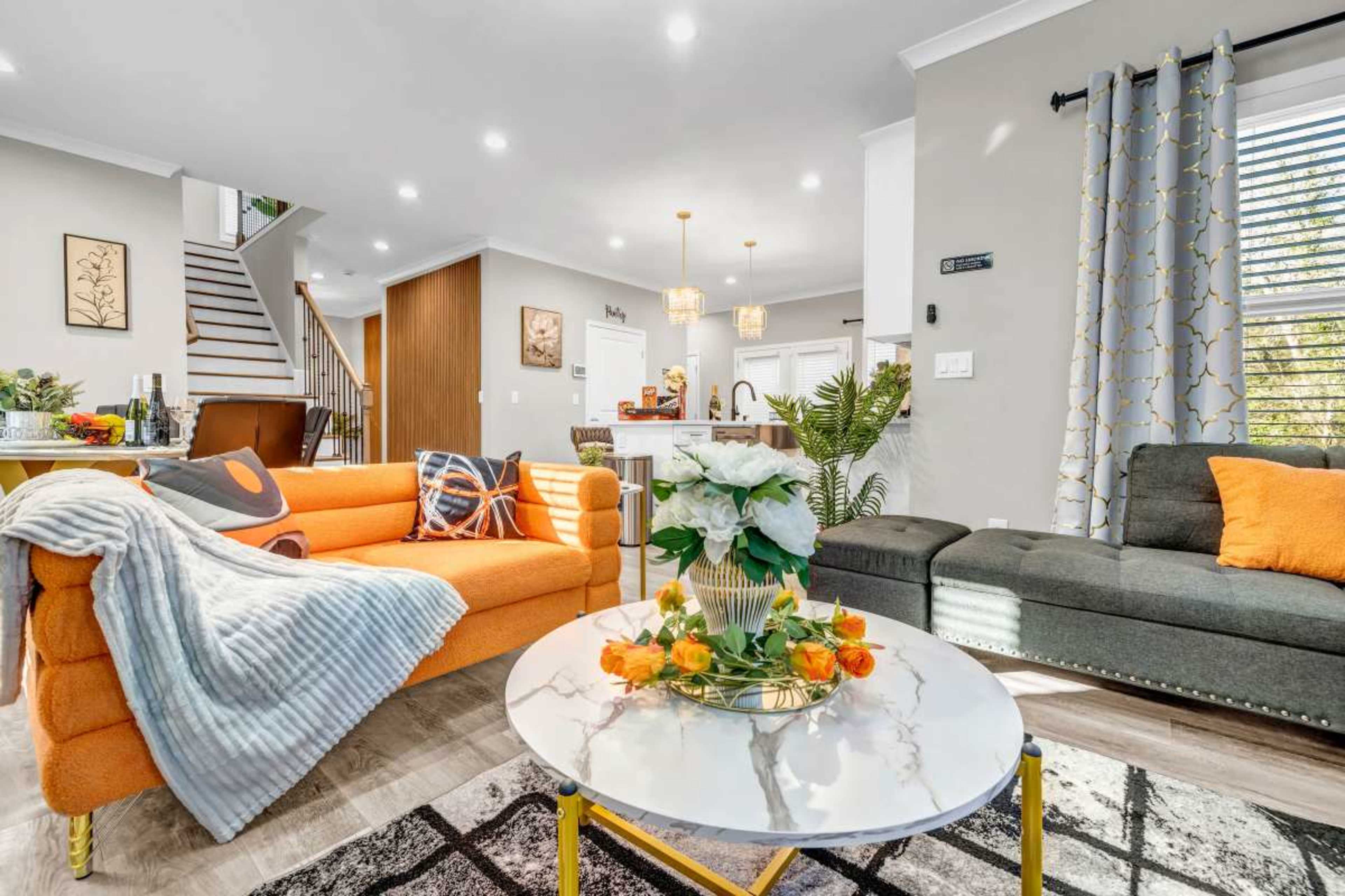 A modern living room features a bright orange couch, a gray sofa, a round coffee table adorned with a floral arrangement, and a staircase in the background.