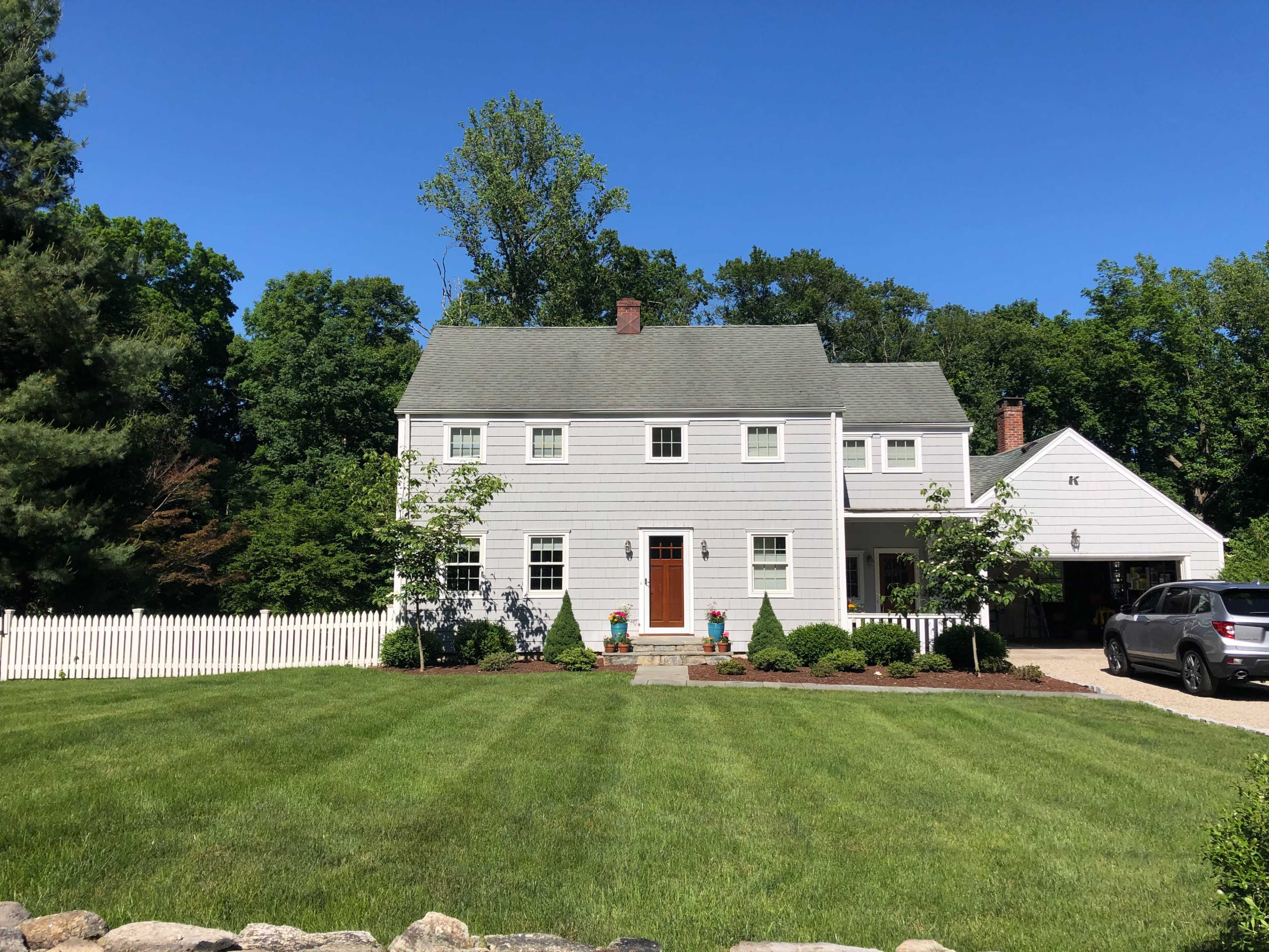 A two-story, light gray house with a front porch and manicured lawn is surrounded by trees and a white picket fence.