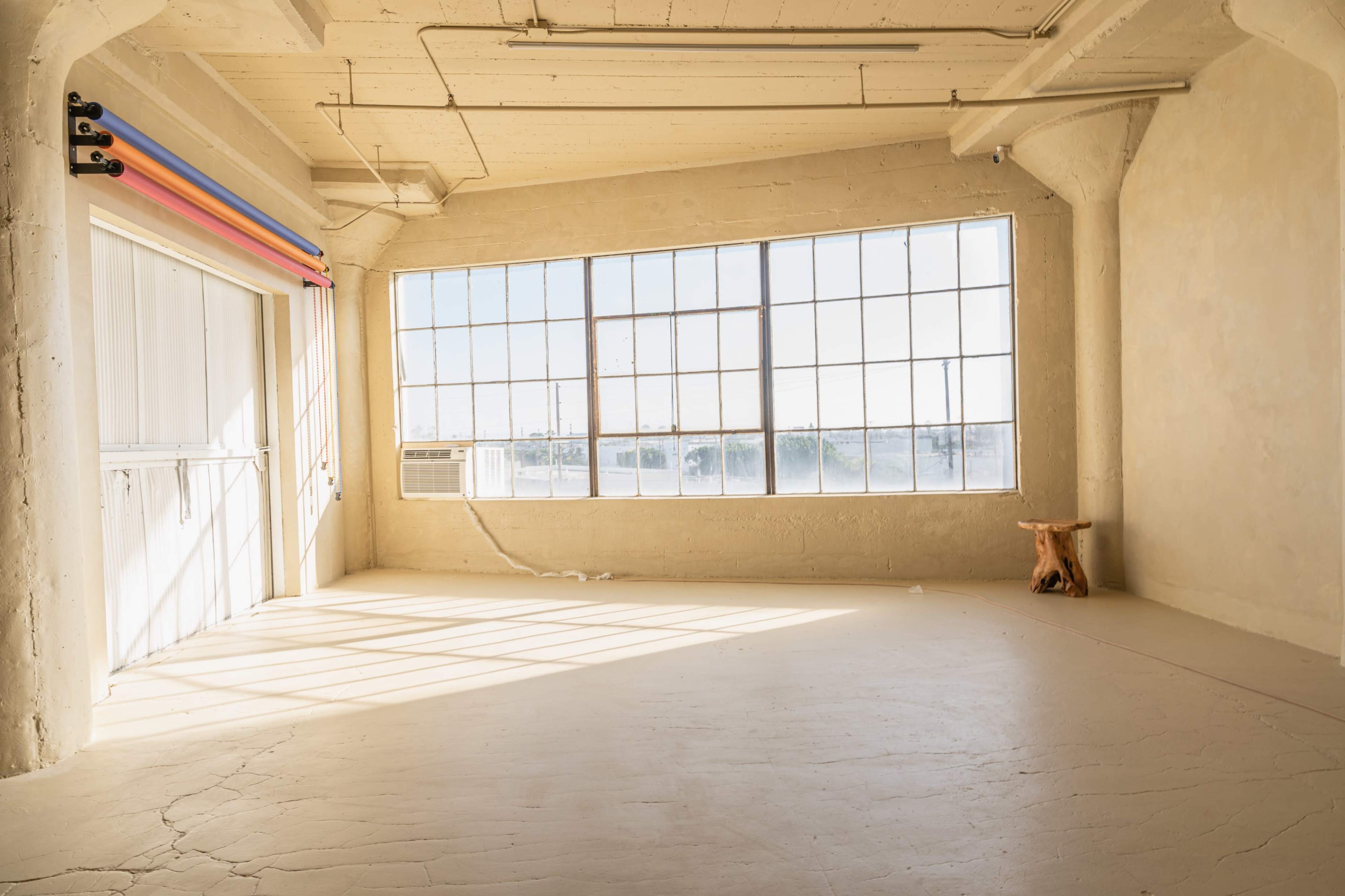 A sunlit room features large windows, bare concrete walls, and a wooden stool in the corner.