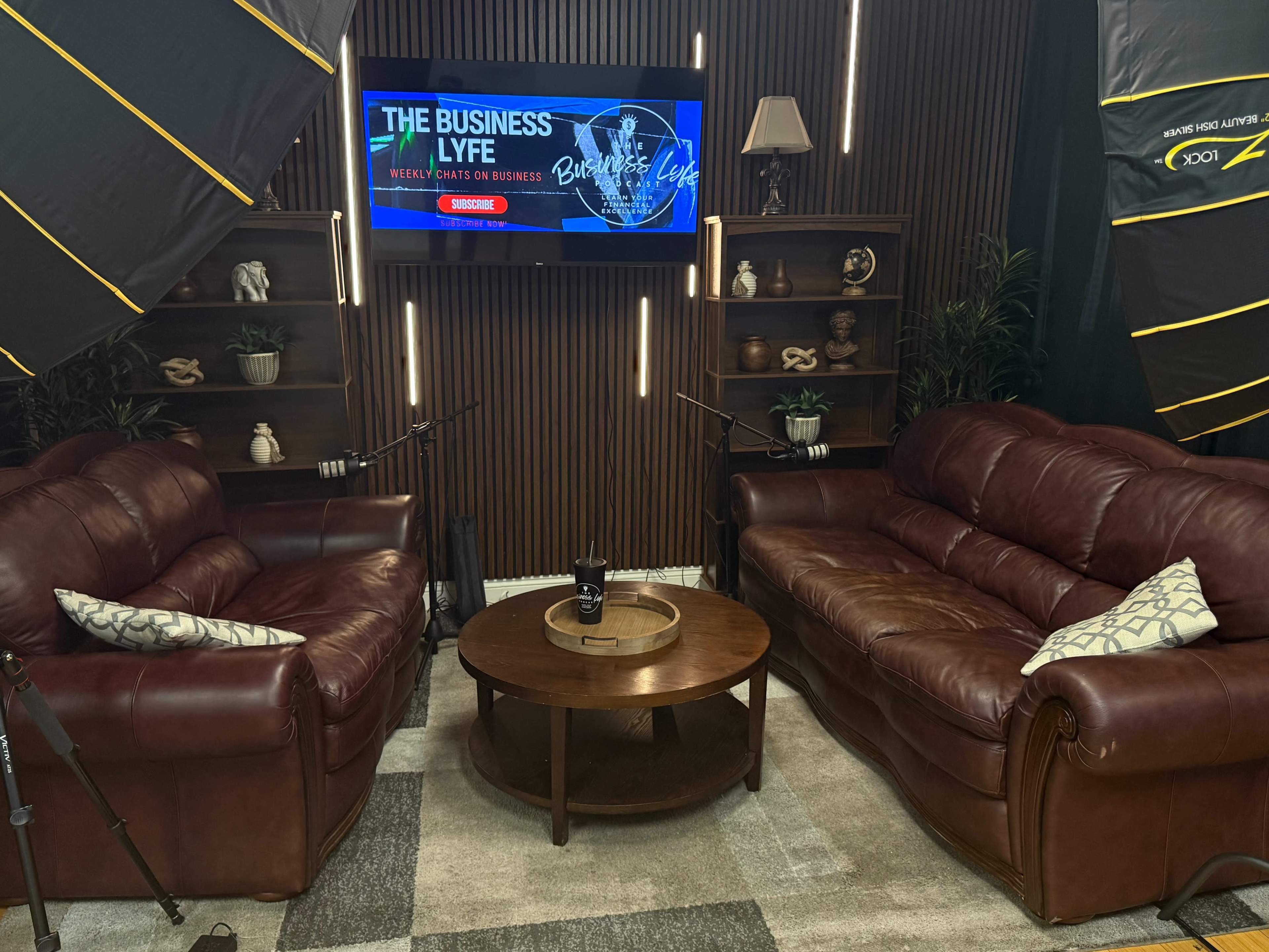 The image shows a studio setup with two brown leather couches facing each other, a round coffee table in the center, and a large screen displaying "THE BUSINESS LYFE" in the background, surrounded by plants and lighting equipment.