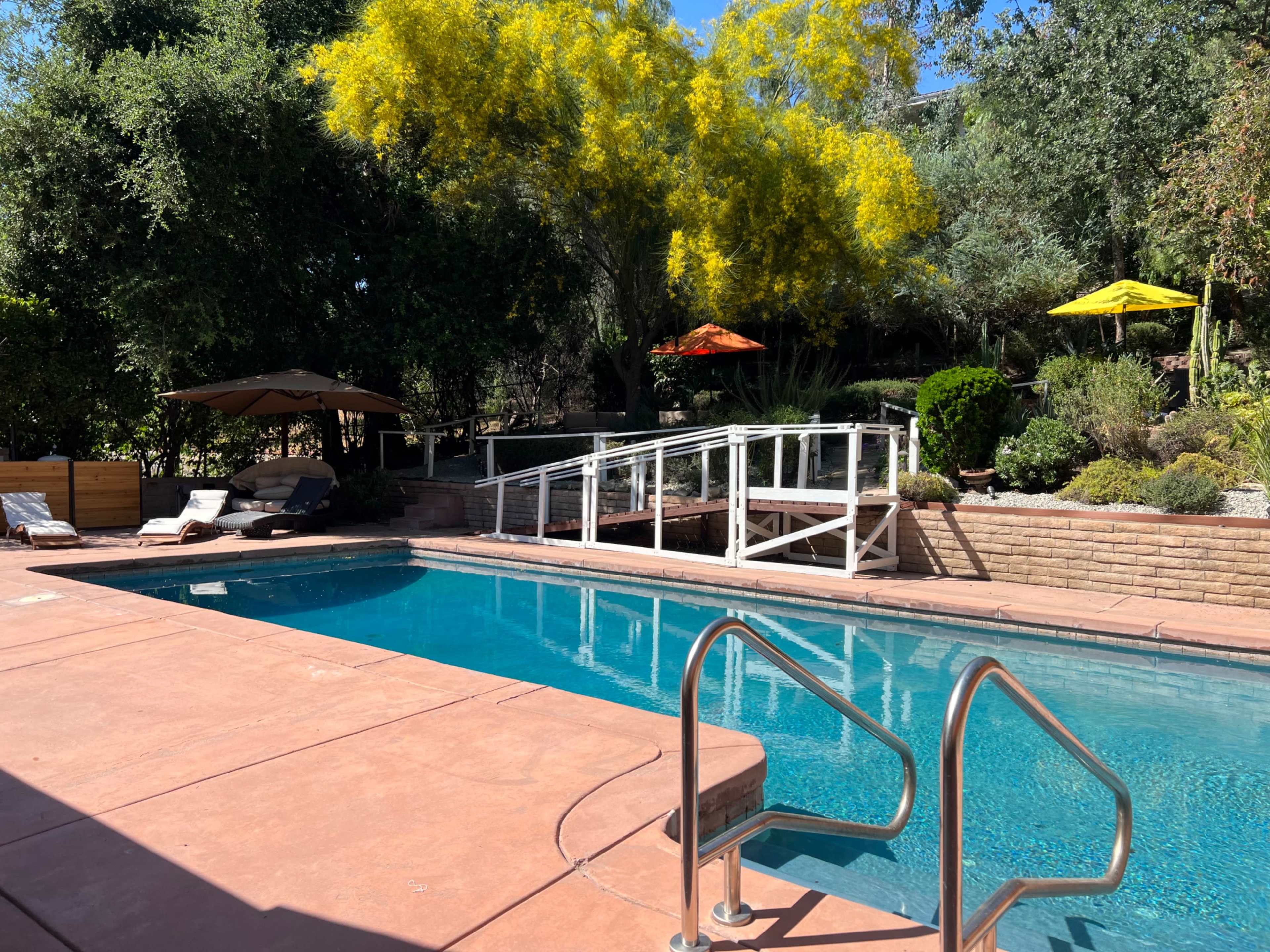 A swimming pool surrounded by a landscaped garden with patio umbrellas and a wooden bridge.