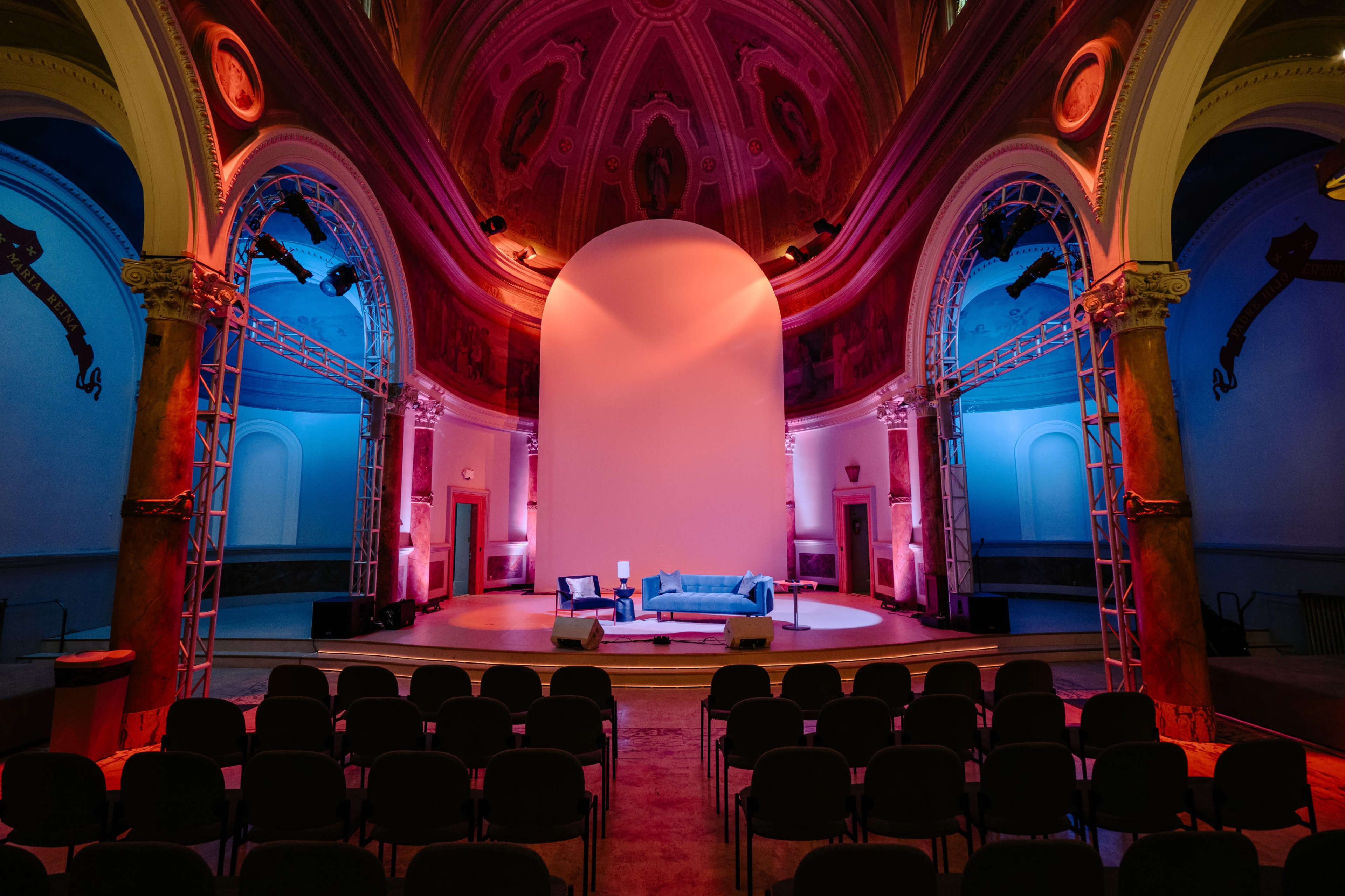 A stage set up in a grand hall with blue and pink lighting, featuring a central white backdrop and a blue couch surrounded by empty rows of chairs.
