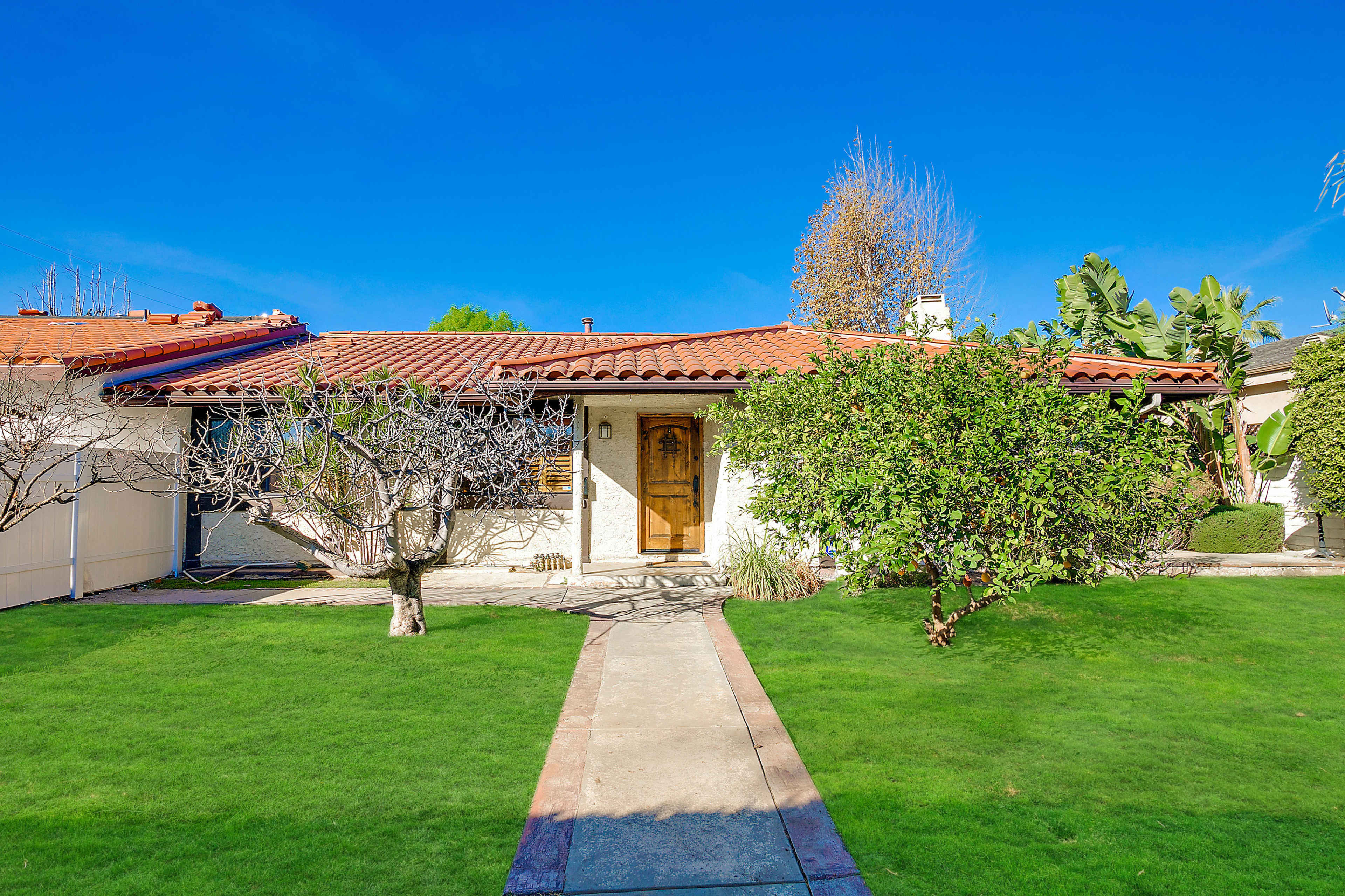 A single-story home with a tiled roof, center front door, and a path leading to it, surrounded by green lawn and landscaped plants.