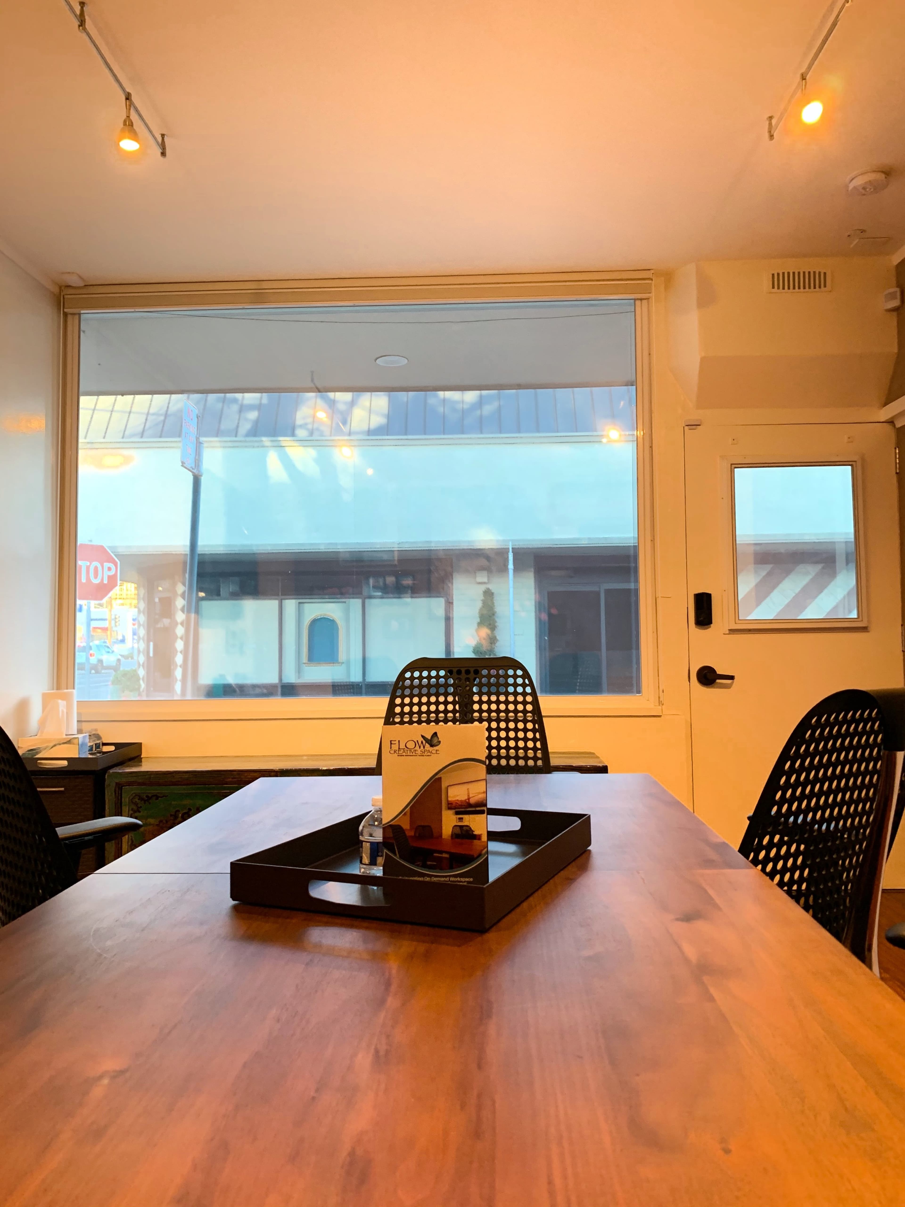 A meeting room features a wooden table with a decorative tray and chairs, surrounded by large windows showcasing a storefront outside.