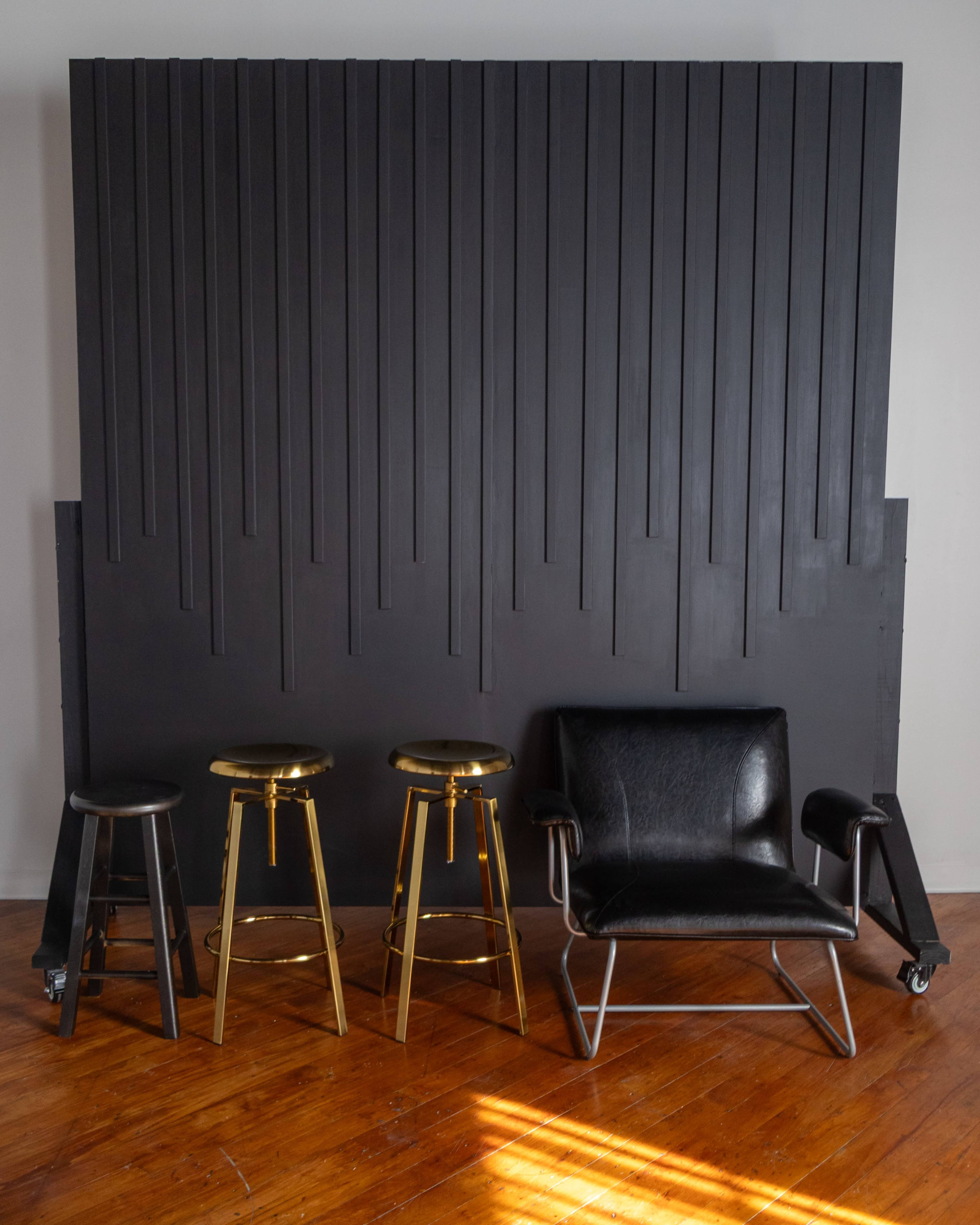 The image features a black wall with vertical grooves, accompanied by three bar stools—two black and one gold—and a black armchair in front of it, all set on a wooden floor.