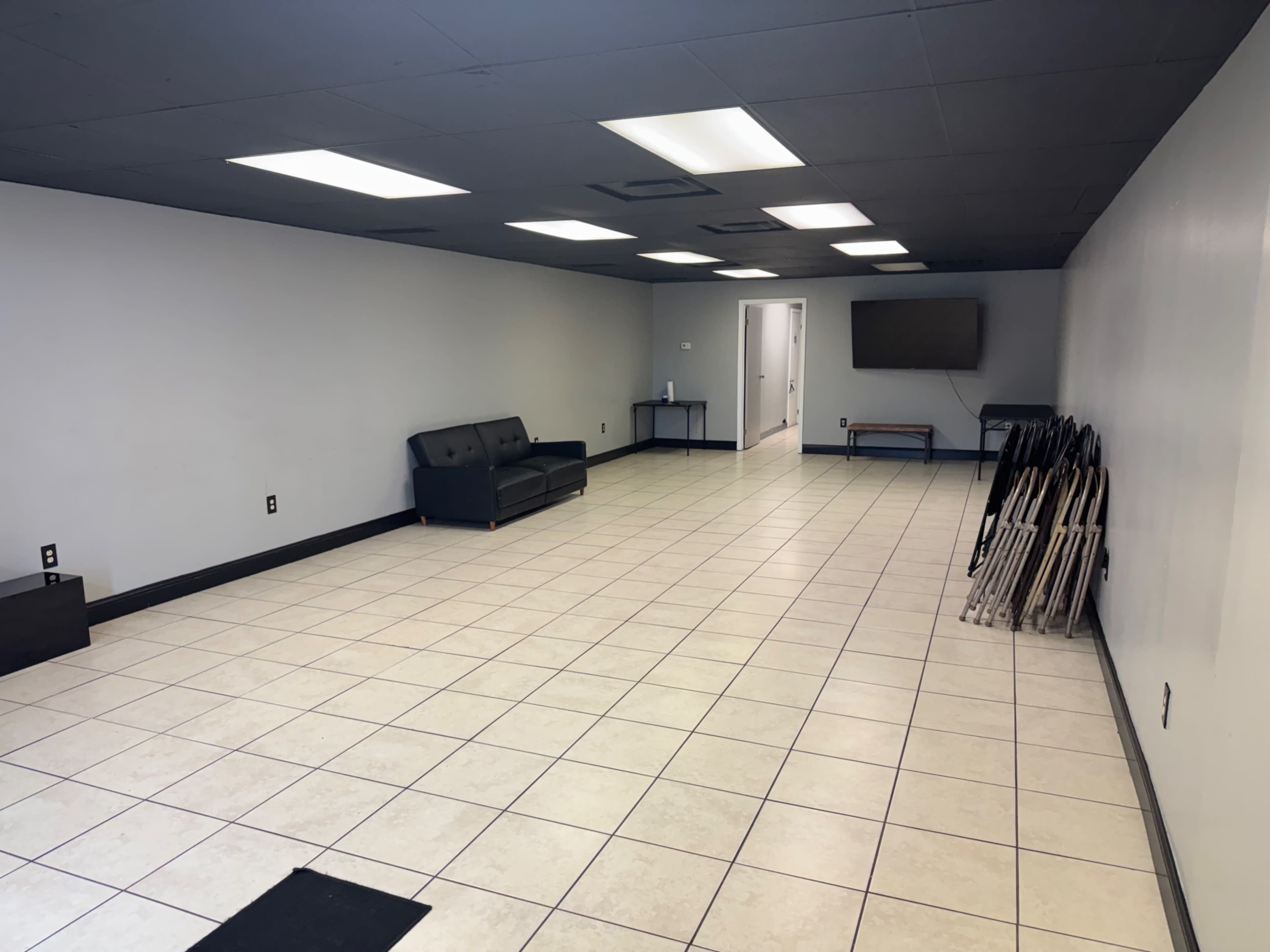 The image shows a spacious, empty room with tiled flooring, a black sofa, and a few folding chairs stacked against the wall.