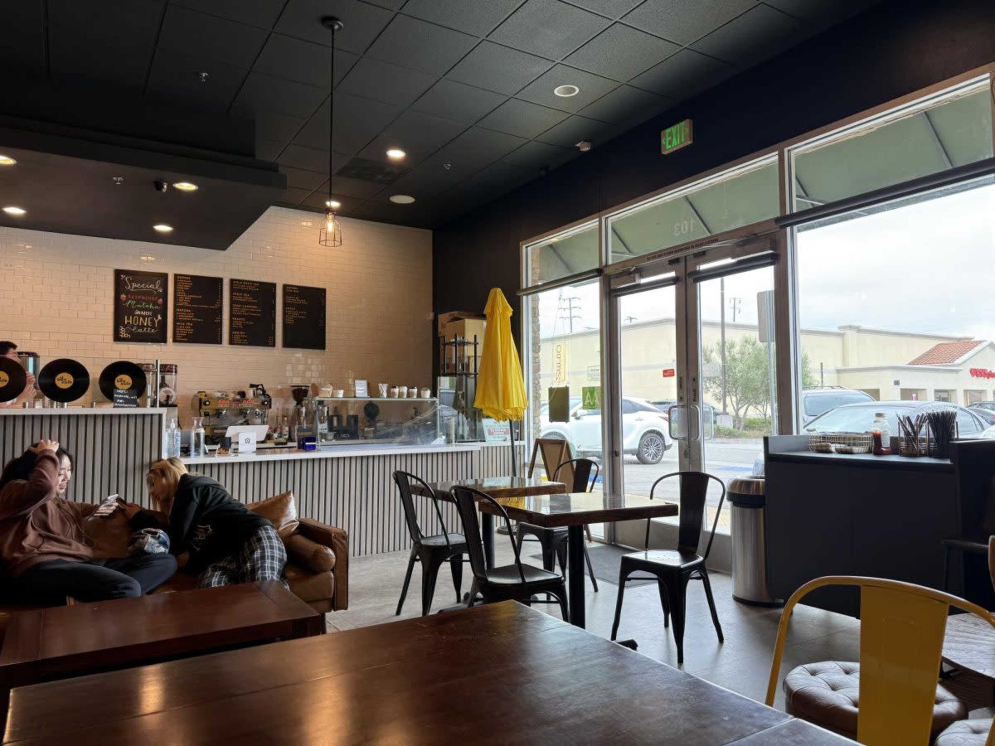The image shows a cozy café interior with wooden tables, black chairs, and a counter displaying a menu, while large windows allow natural light to enter.