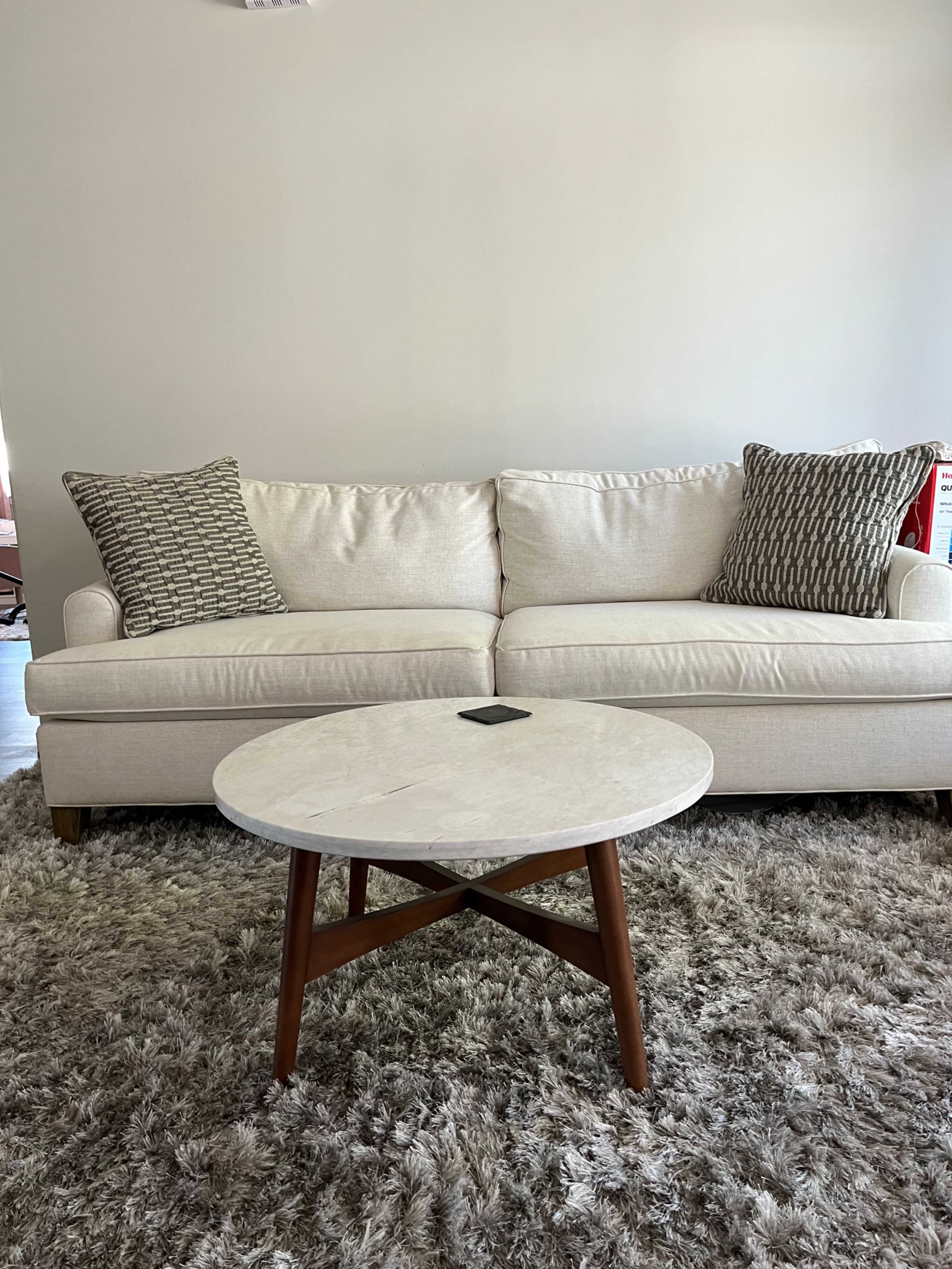 A light-colored sofa with patterned cushions is positioned in front of a circular marble-topped coffee table on a textured rug.