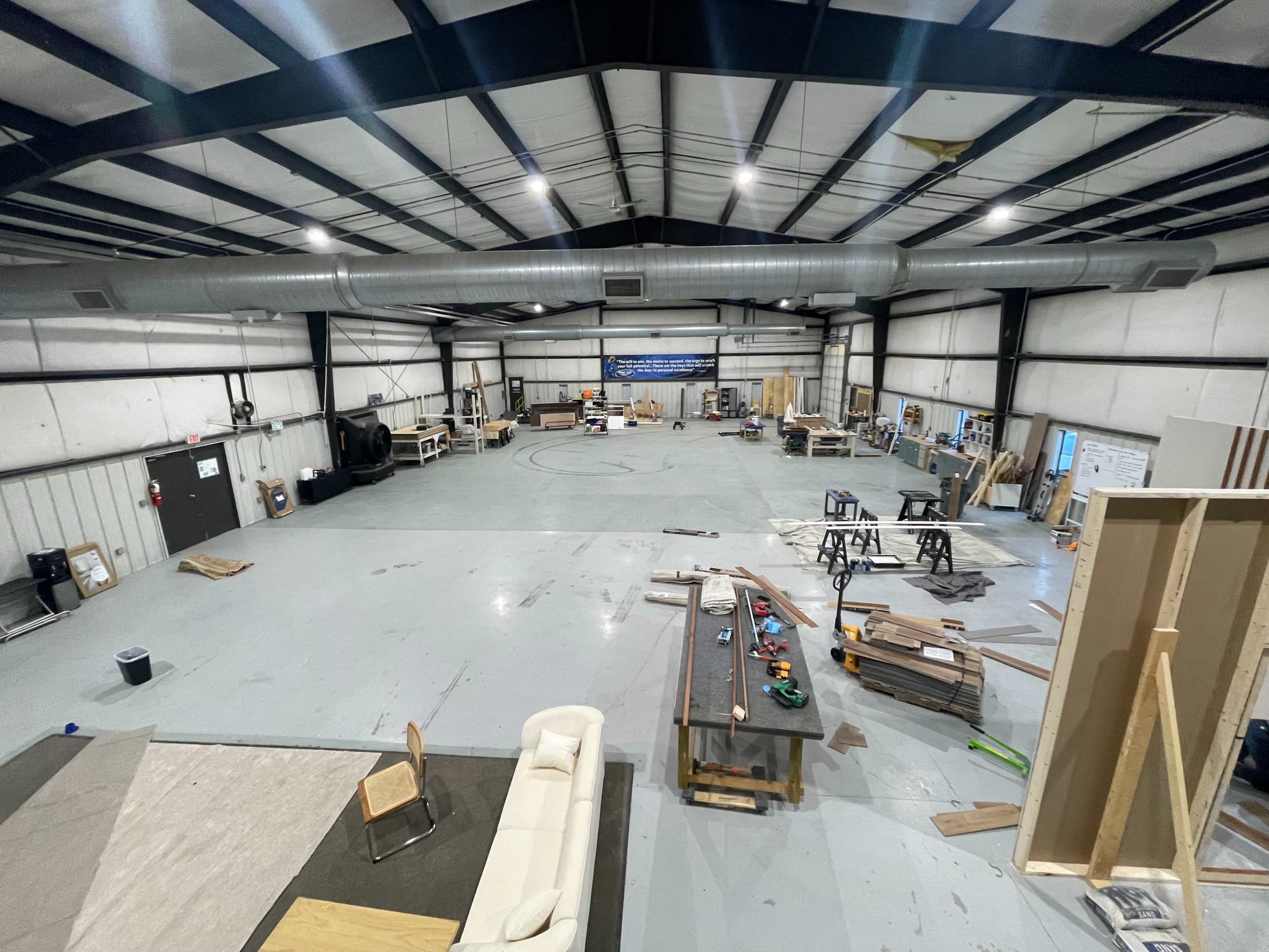 The image shows a spacious industrial warehouse filled with various workstations, tools, and materials, organized for construction or crafting activities.