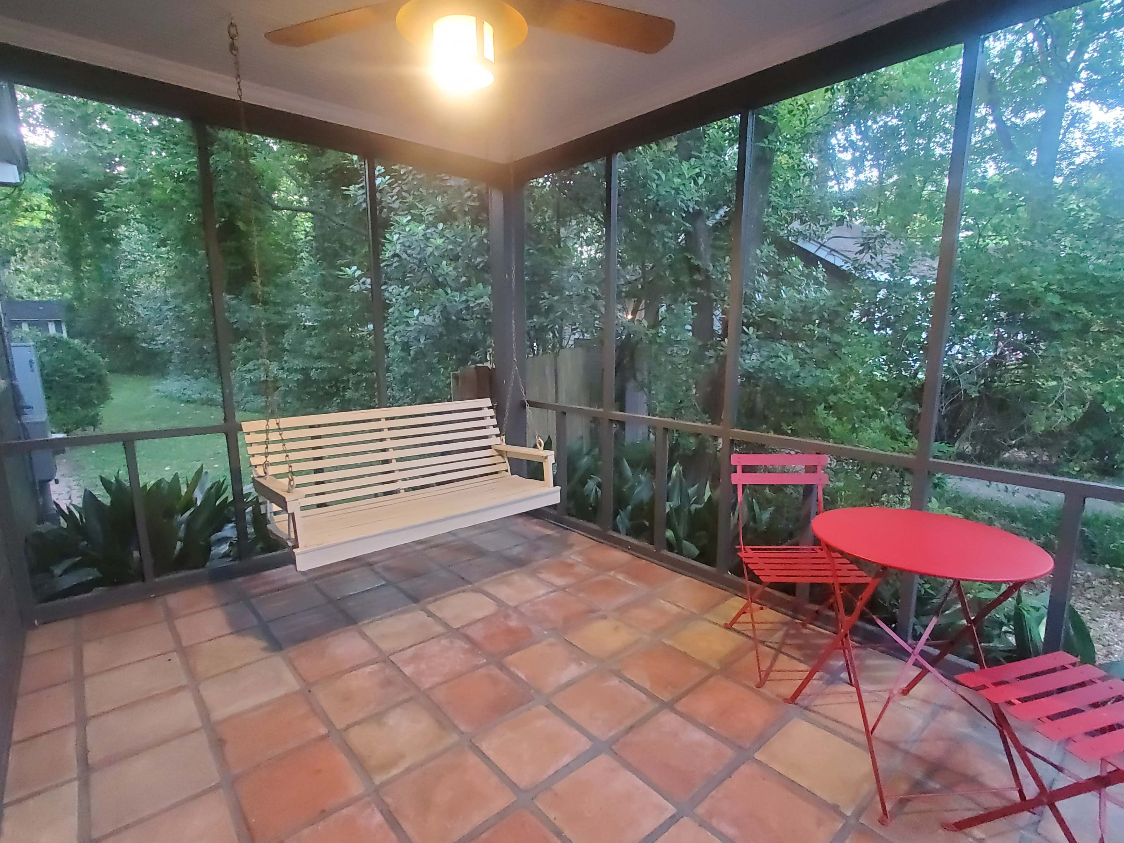 The image shows a screened porch with a wooden swing and a small red table with two matching chairs, surrounded by greenery.