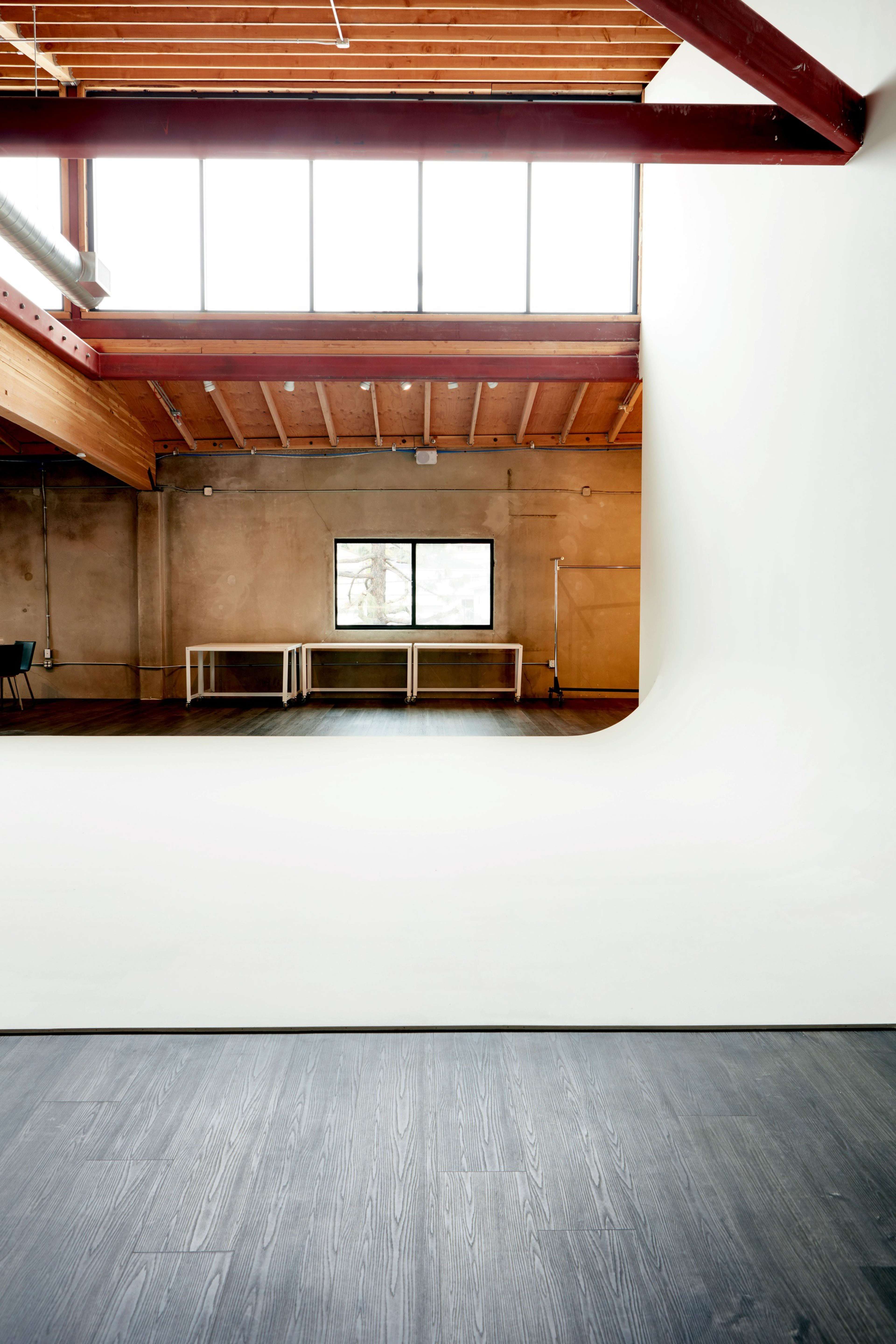 An empty studio space with wooden beams, a large white backdrop, and a window overlooking the interior.