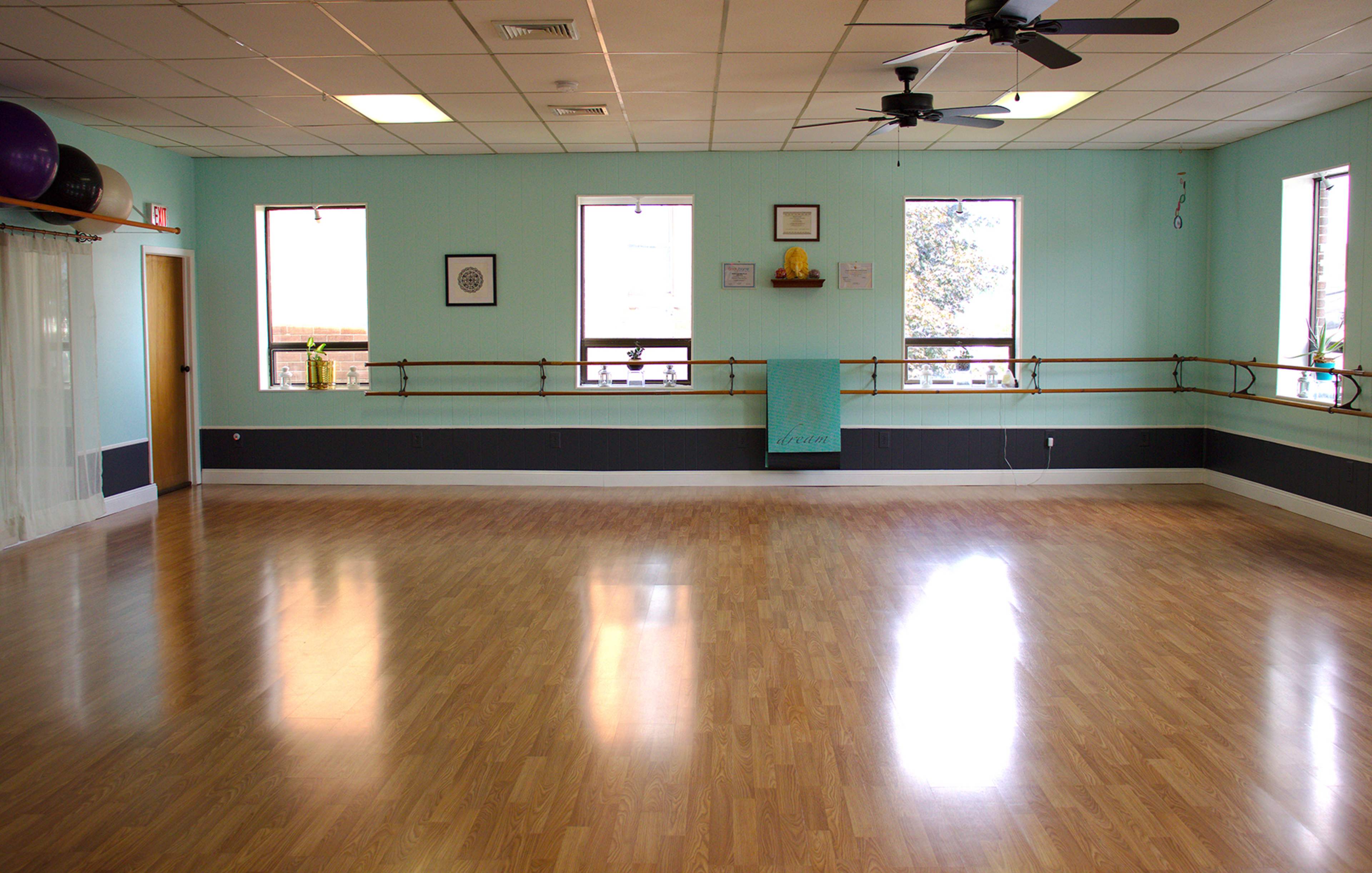 A spacious dance studio with wooden flooring, large windows, and a ballet barre along the walls.