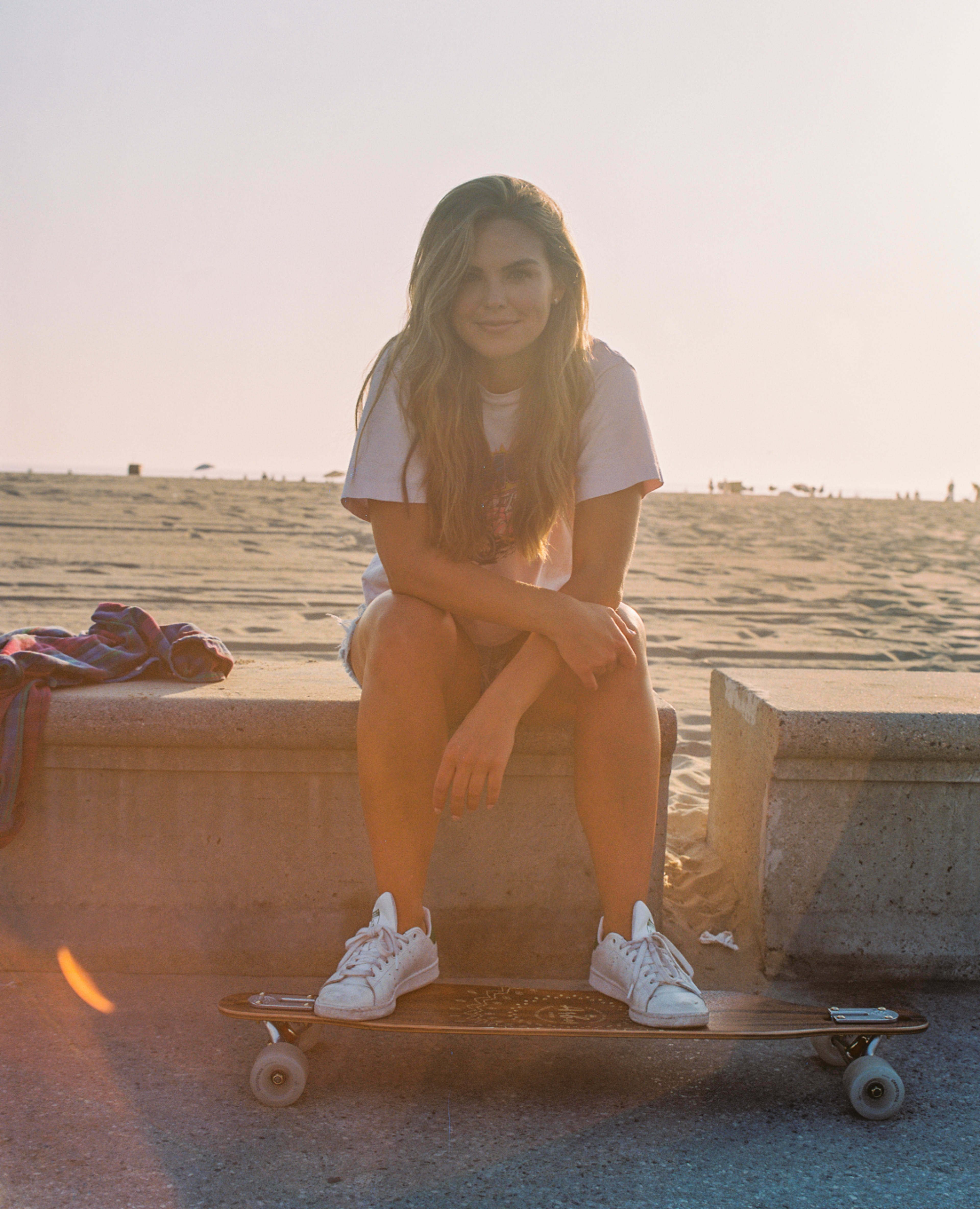 A young woman sits on a concrete barrier with a skateboard at her feet, smiling in the warm glow of sunset on a beach.