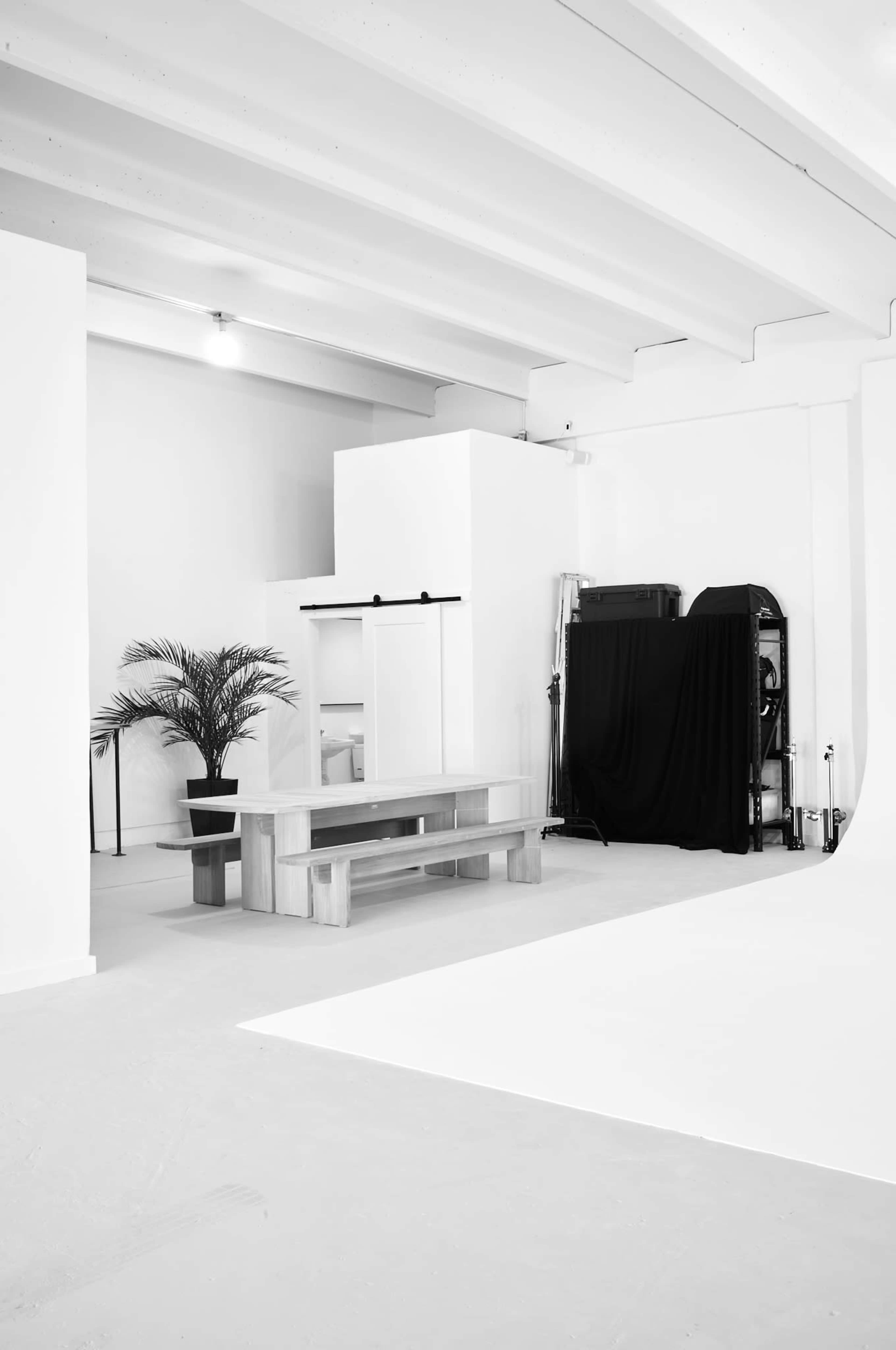 A minimalist studio space featuring a wooden table, a potted plant, and various equipment against stark white walls.
