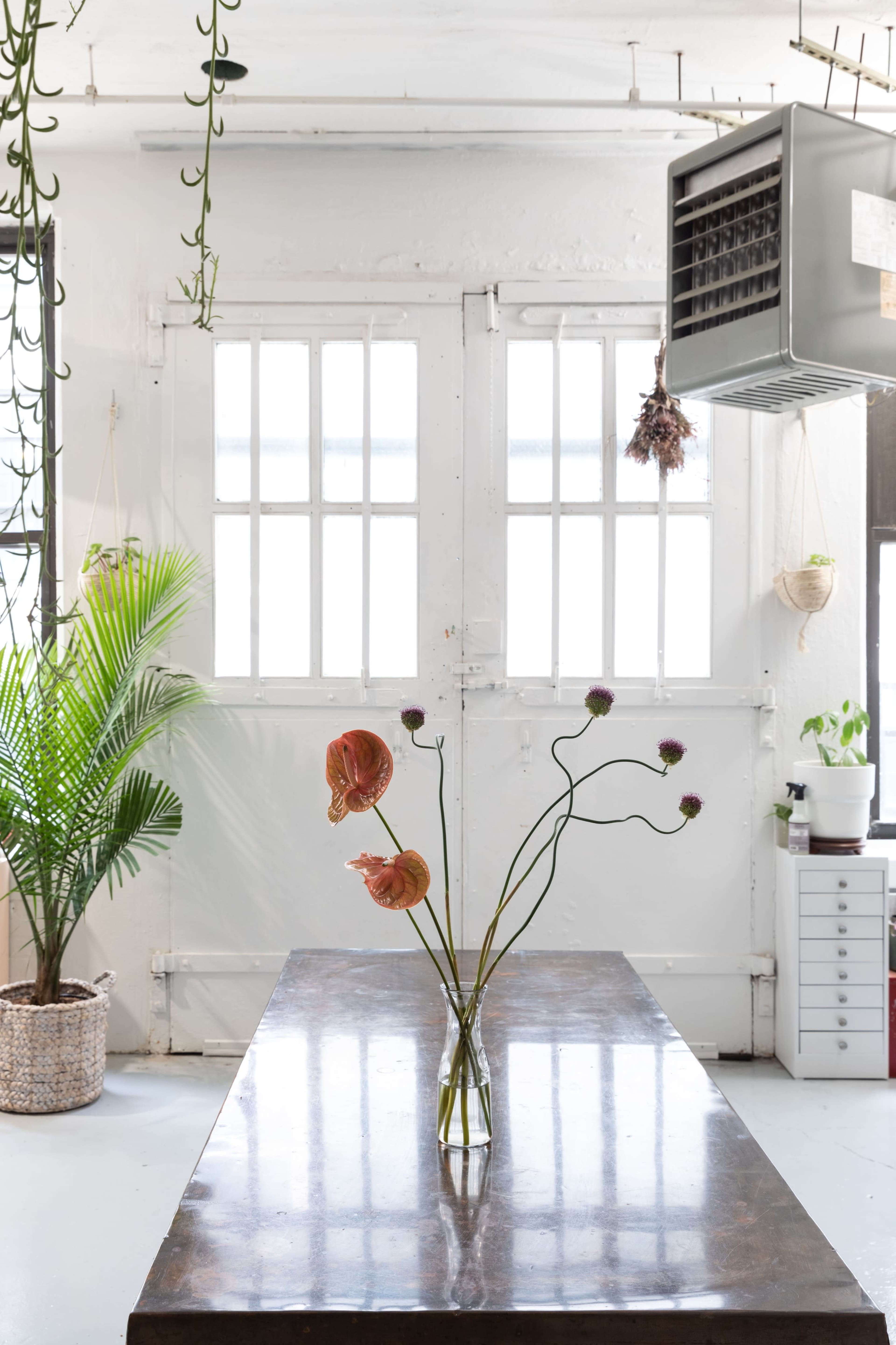 A polished table displays a vase with flowers, while large windows and plants create a bright, open space.