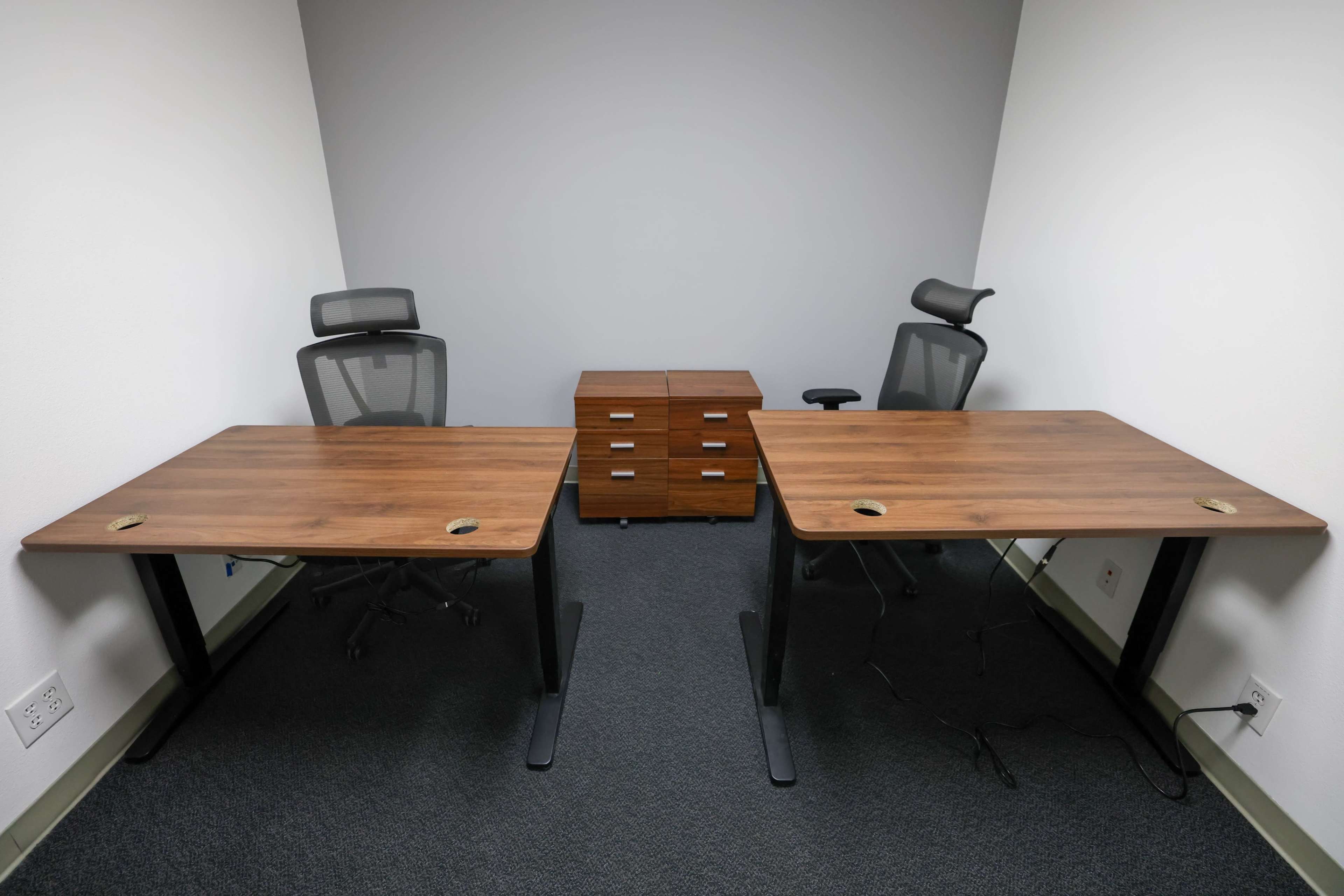 Two wooden desks with ergonomic chairs positioned in an empty office space, accompanied by a chest of drawers.