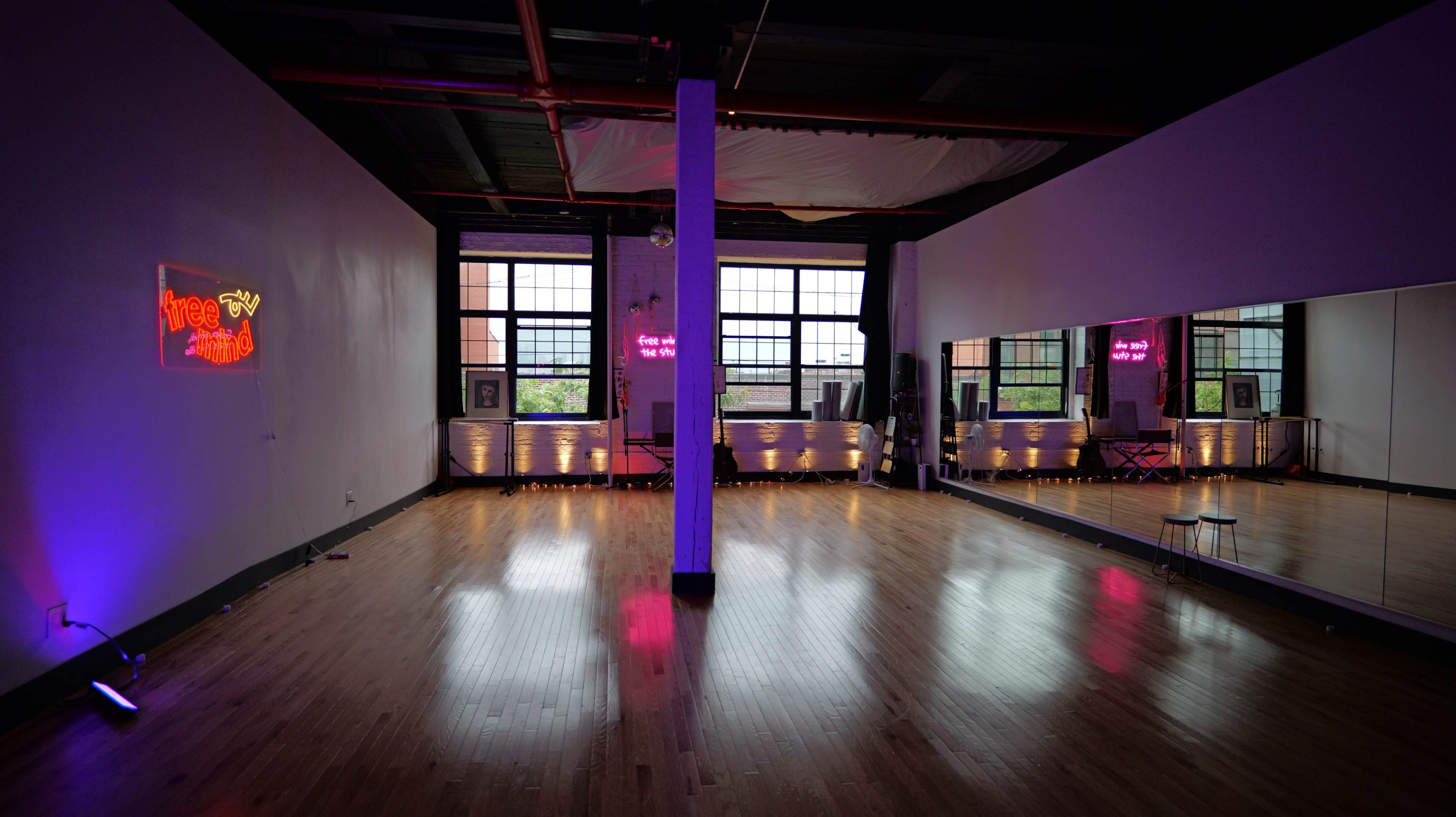 A spacious dance studio with hardwood floors, large windows, and neon signs illuminating the walls.
