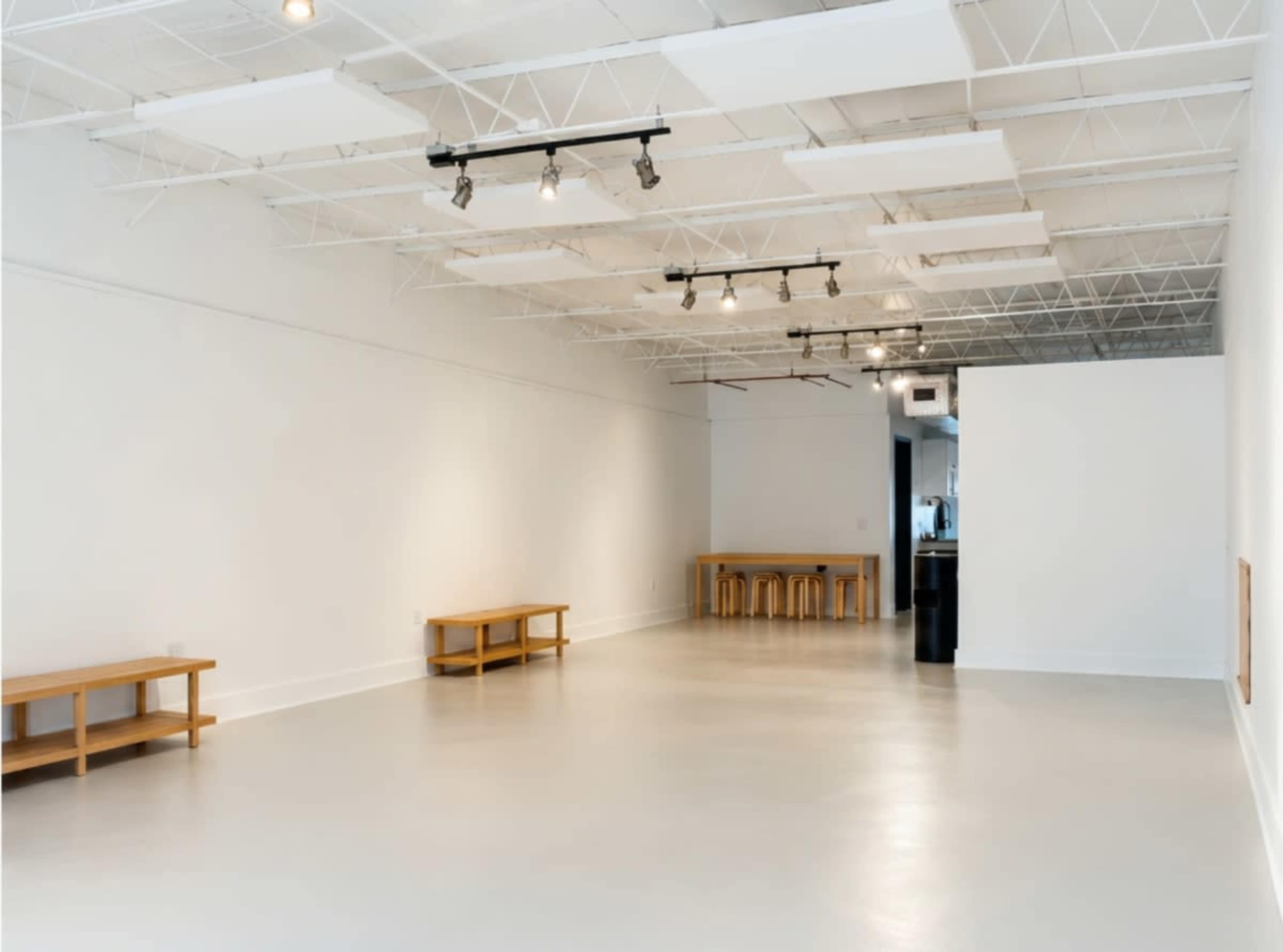 The image shows a spacious, empty room with white walls, a polished concrete floor, and minimal wooden furniture along the sides.