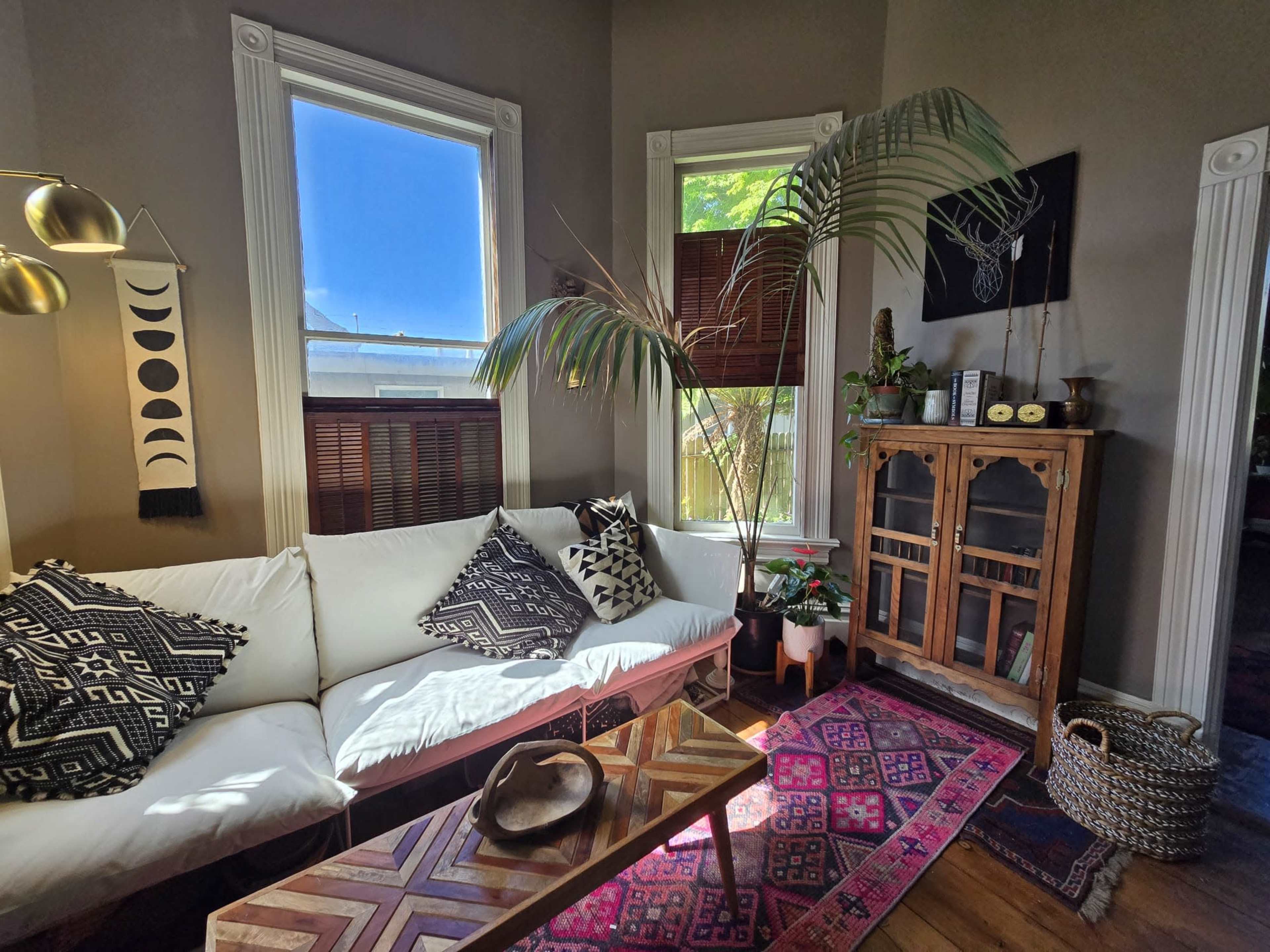 The image shows a cozy living room with a white couch, patterned cushions, a wooden coffee table, and a decorative cabinet beside a large potted plant.