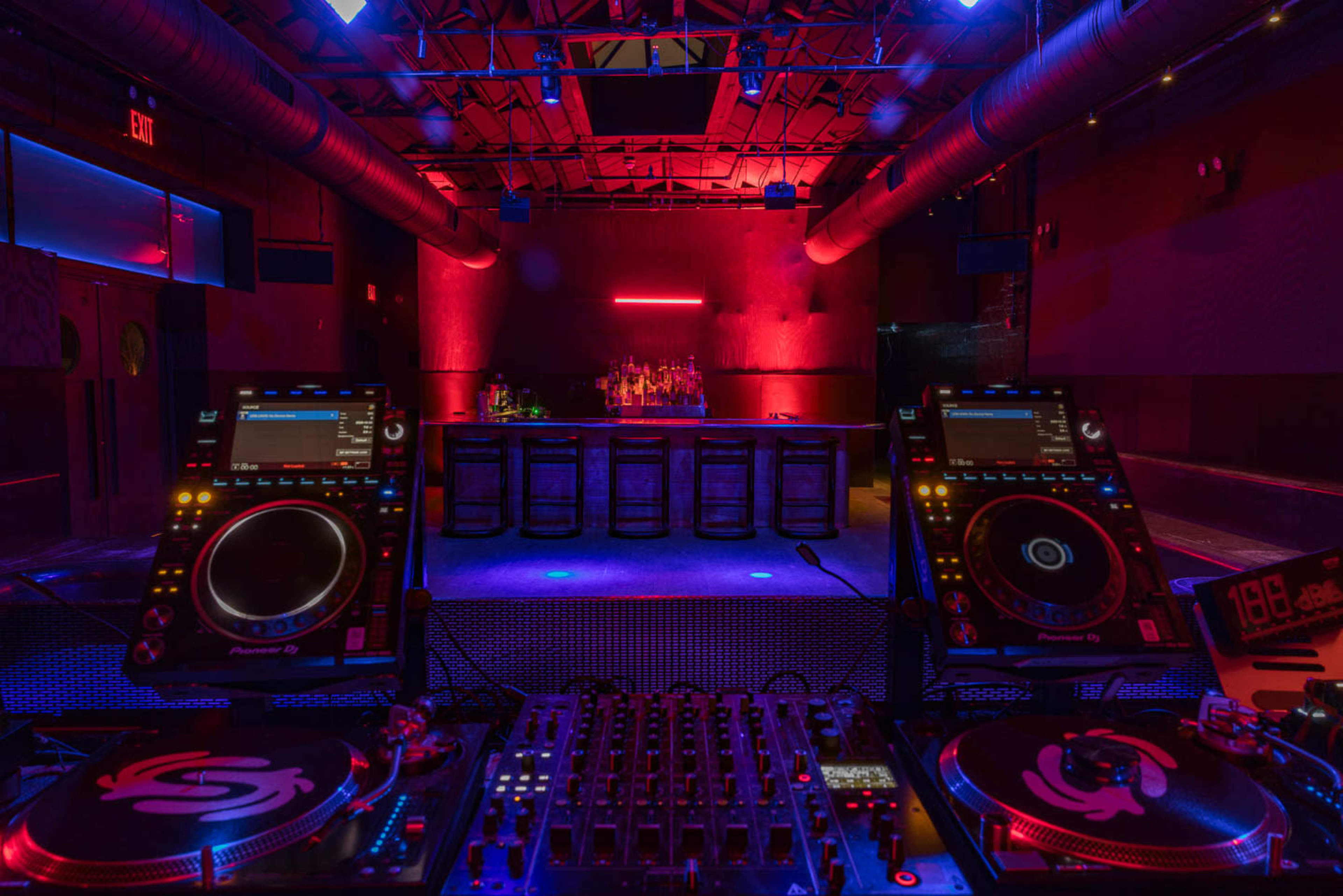 A dimly lit nightclub features DJ equipment in the foreground and a bar with illuminated bottles in the background.