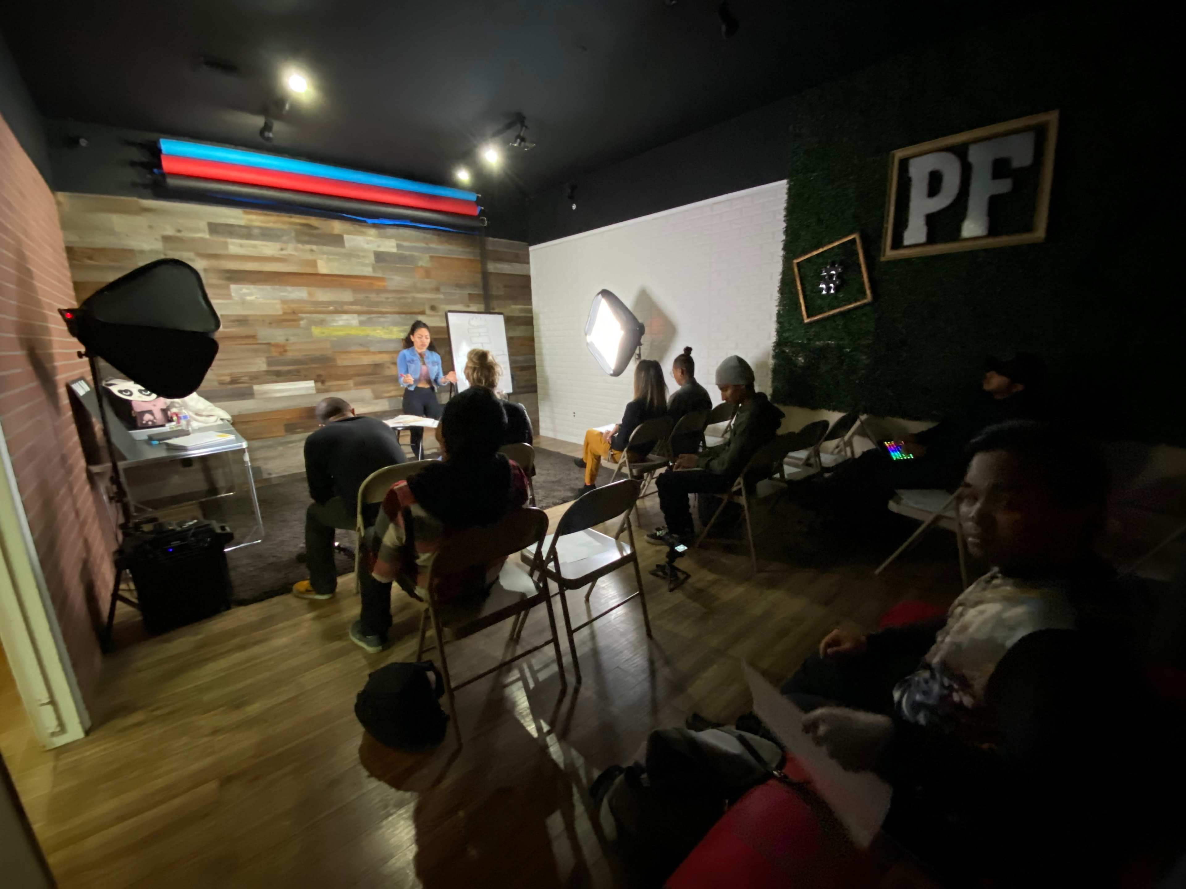 A group of people is seated in a room with wooden walls, listening to a presentation from a woman standing in front of a whiteboard.