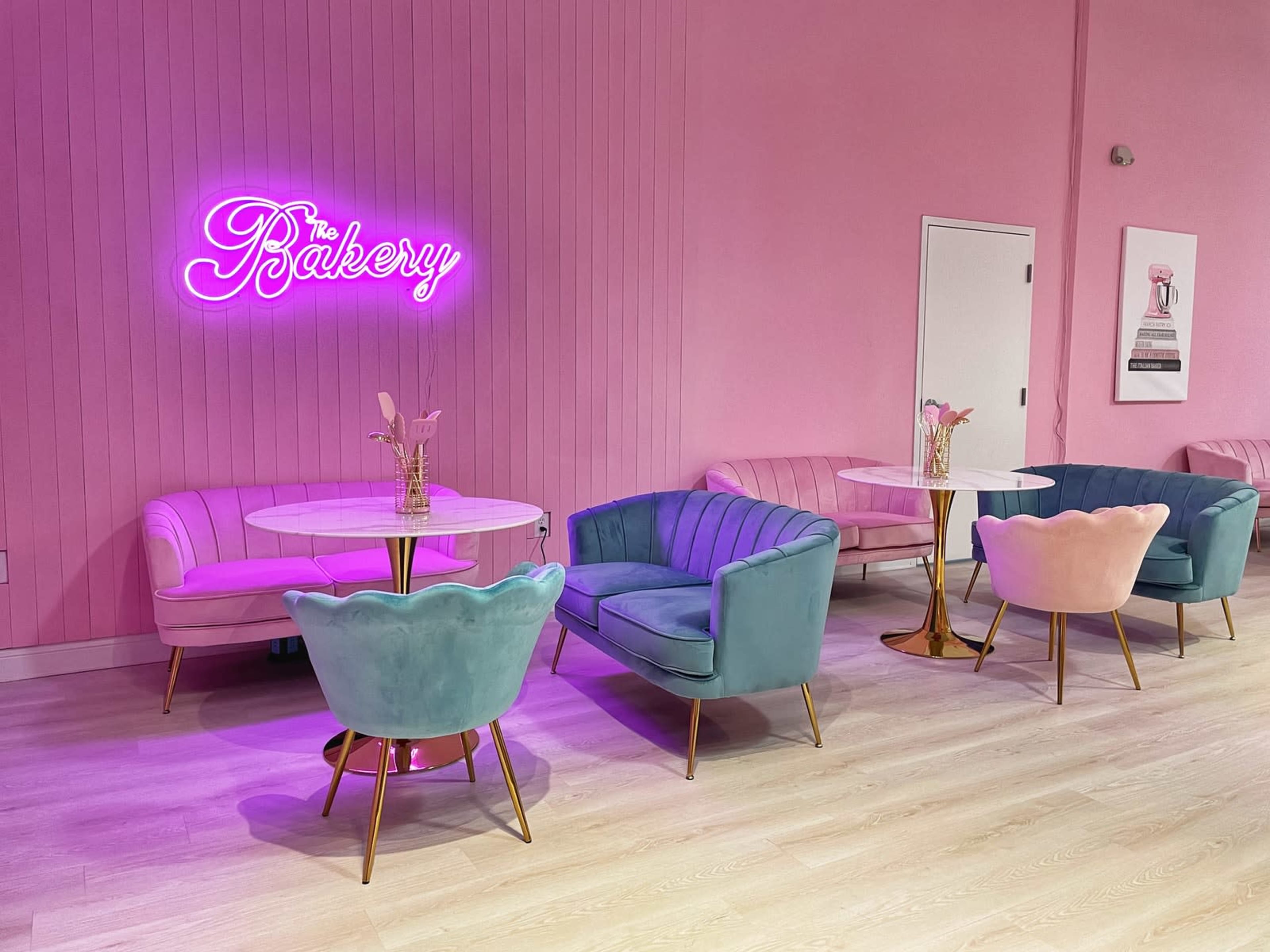 A stylish café interior with pastel-colored furniture, including pink and blue chairs, round tables, and a neon sign that says "The Bakery" on a pink wall.