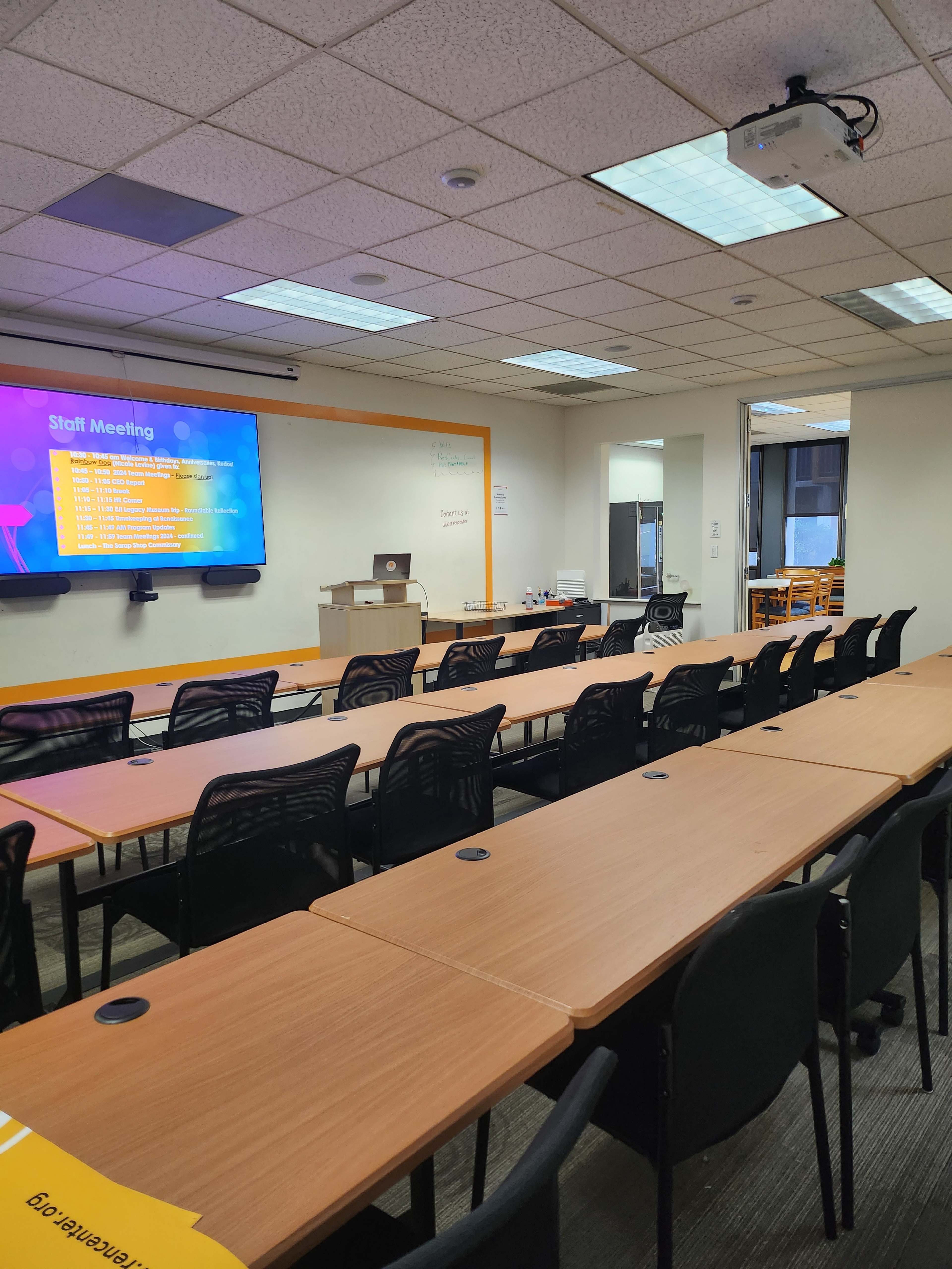 A conference room is set up with rows of tables and chairs facing a projector screen displaying a meeting agenda.