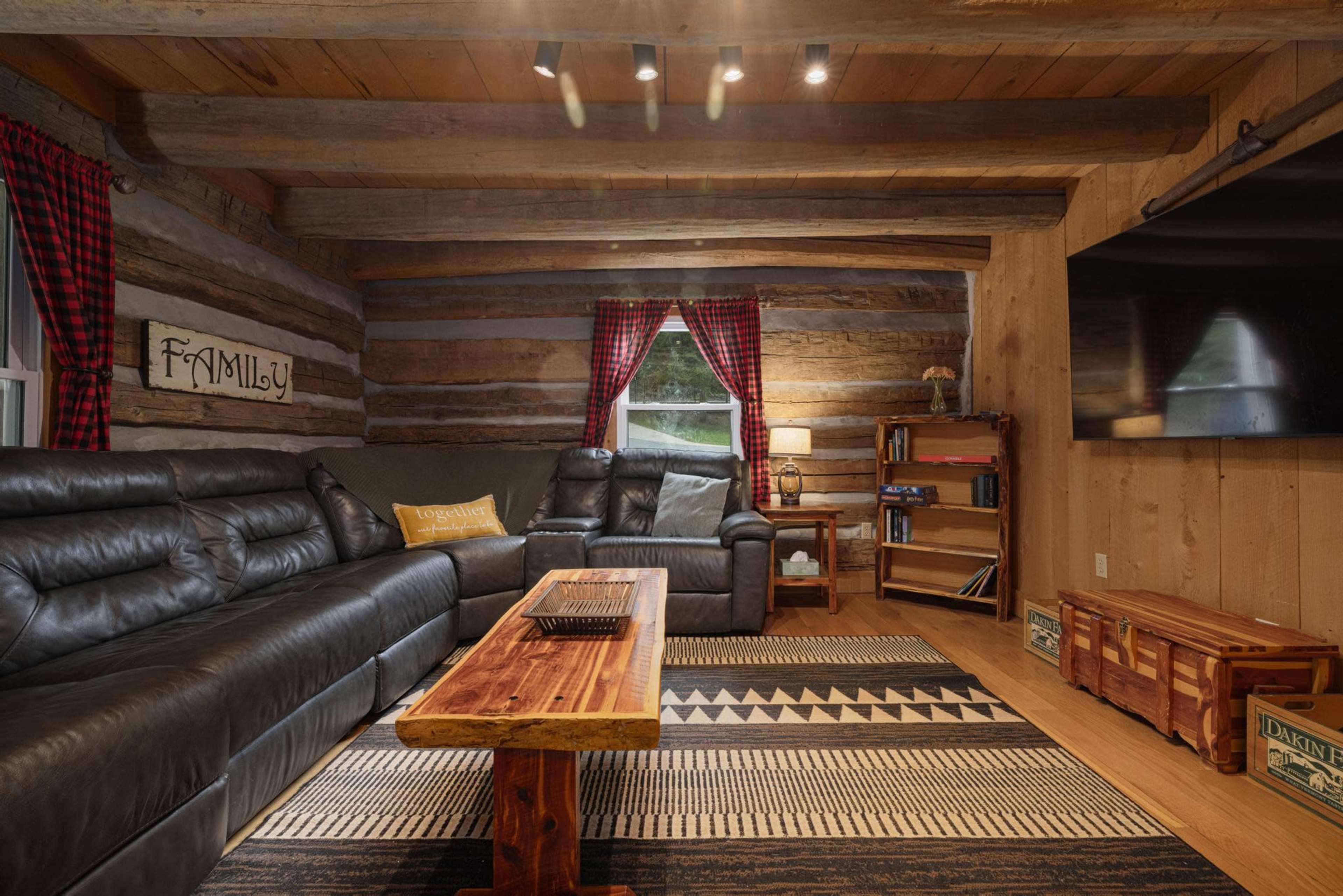 The image shows a cozy living room with log walls, a large leather sectional sofa, a wooden coffee table, and a flat-screen television mounted on one wall.