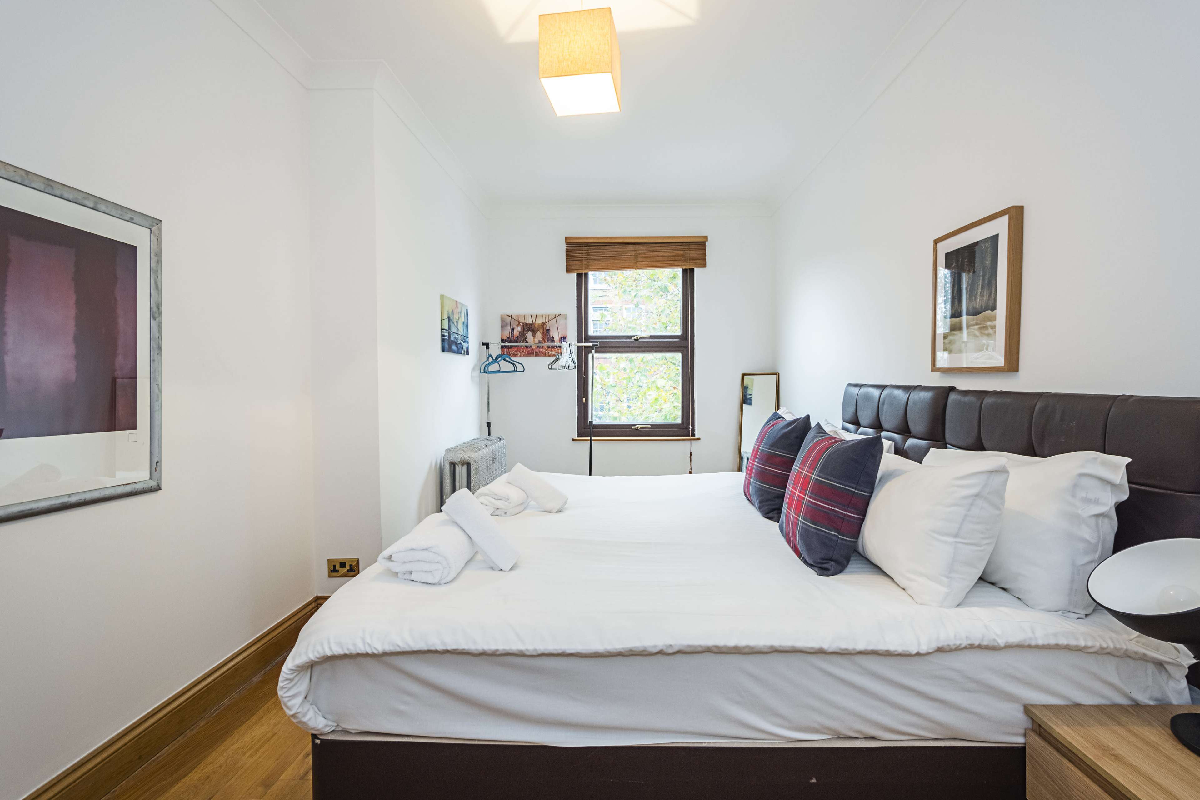 The image shows a small, neatly arranged bedroom with a double bed, two pillows, and a window offering natural light.
