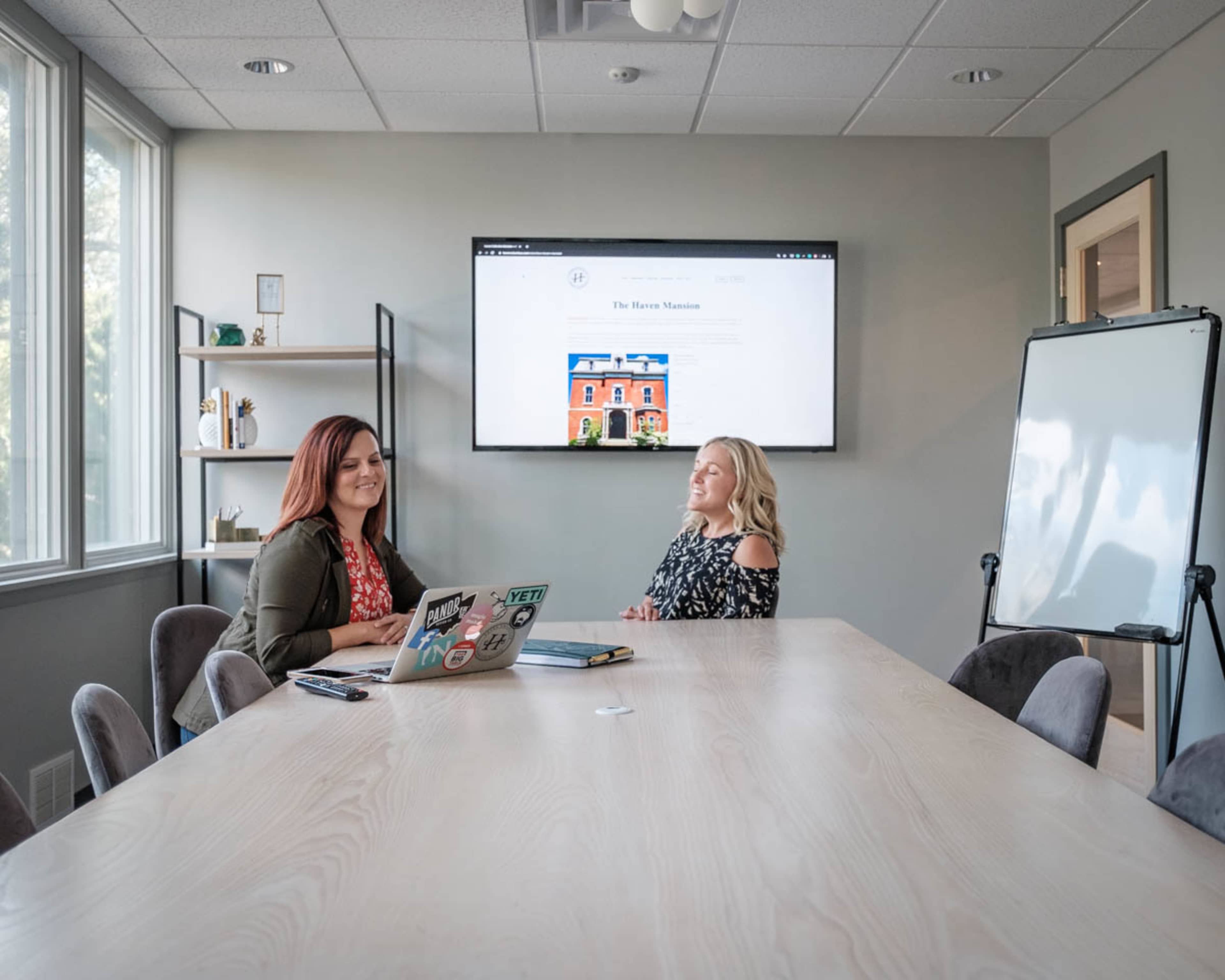 Two women sit at a conference table in a meeting room, with a wall-mounted screen displaying a website in the background.