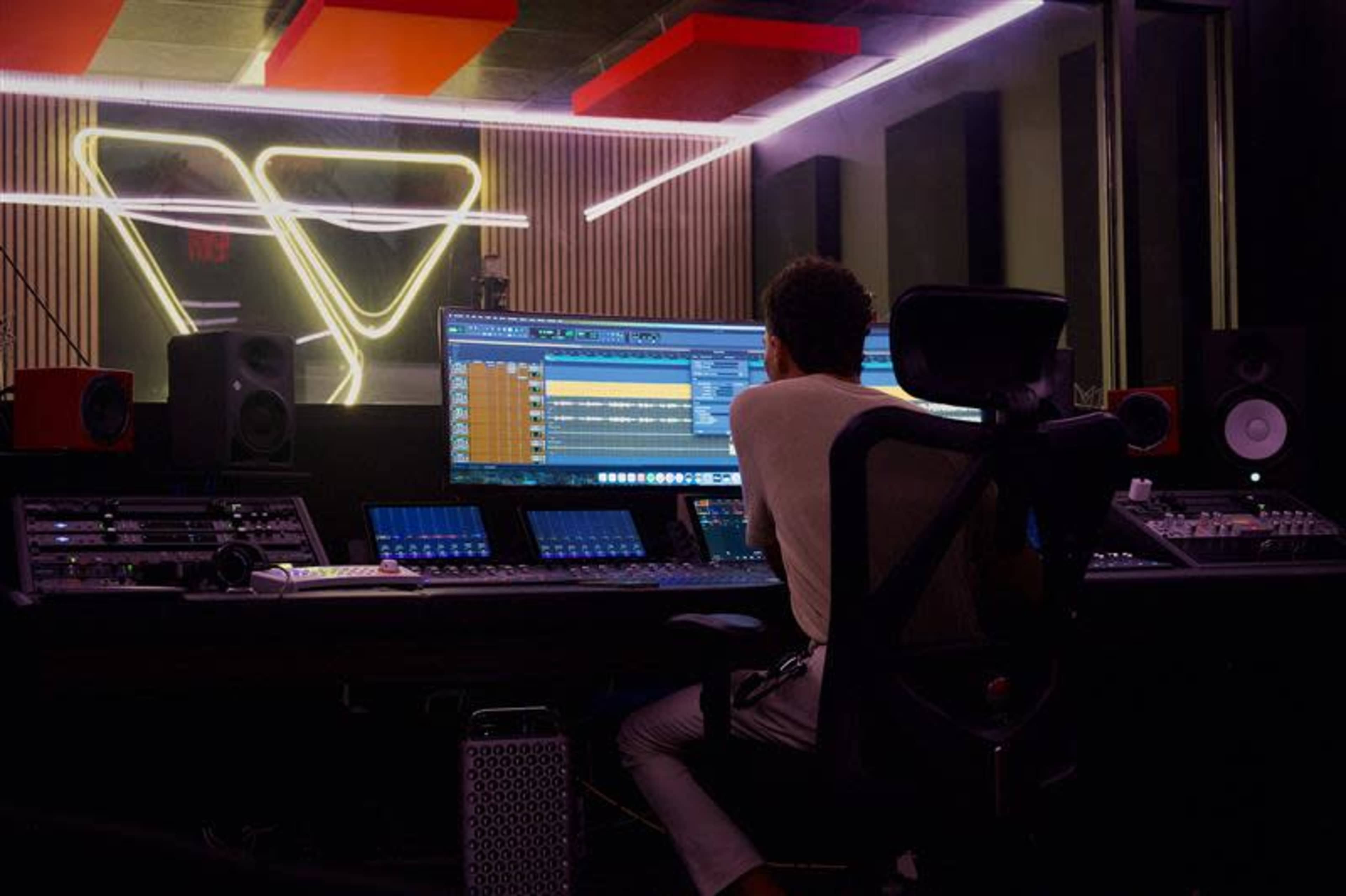A person is working at a large mixing console in a music studio, analyzing audio on a computer screen surrounded by various sound equipment.