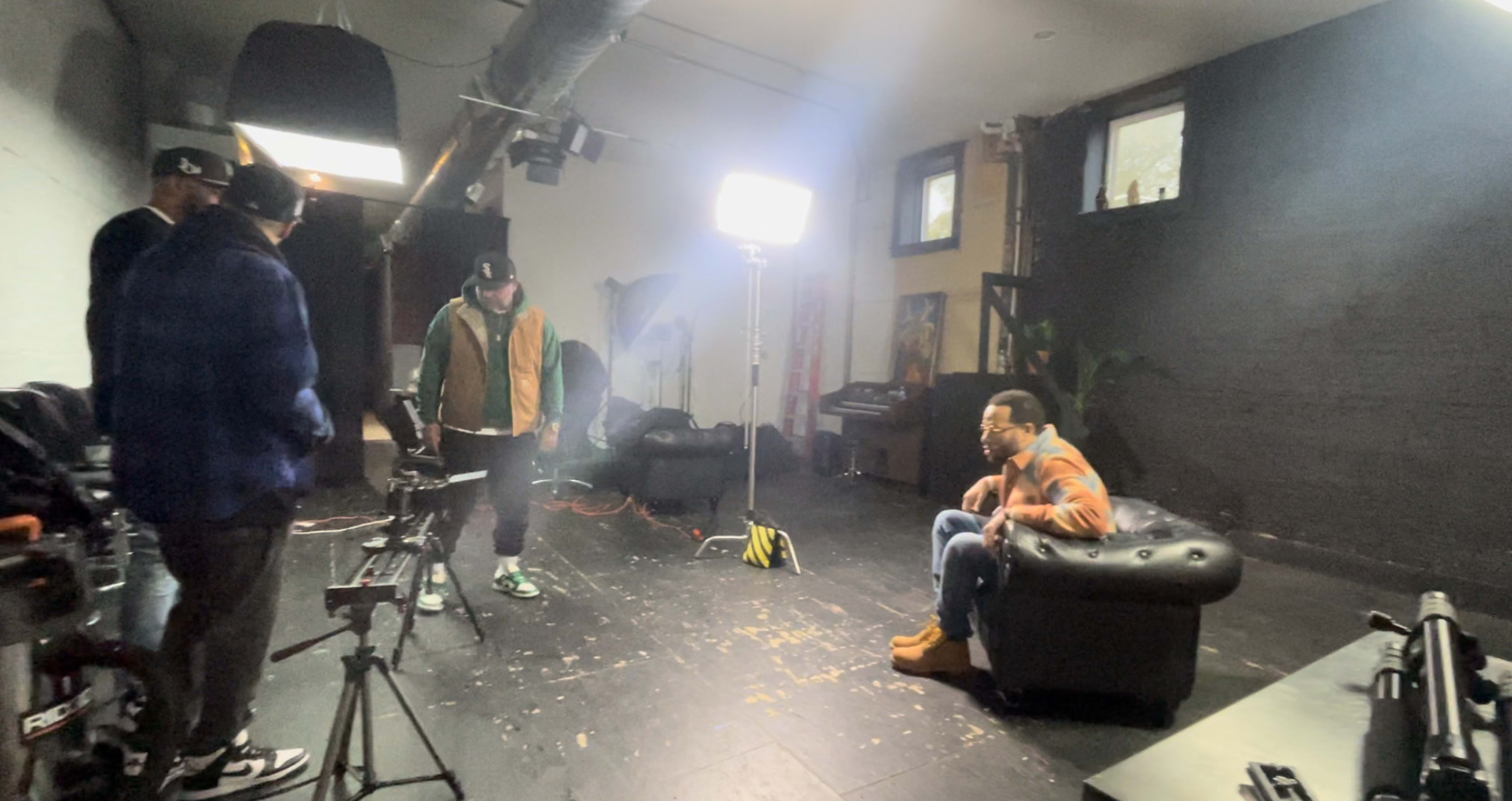 A group of three people is in a dimly lit studio setting with lighting equipment, cameras, and a black couch.