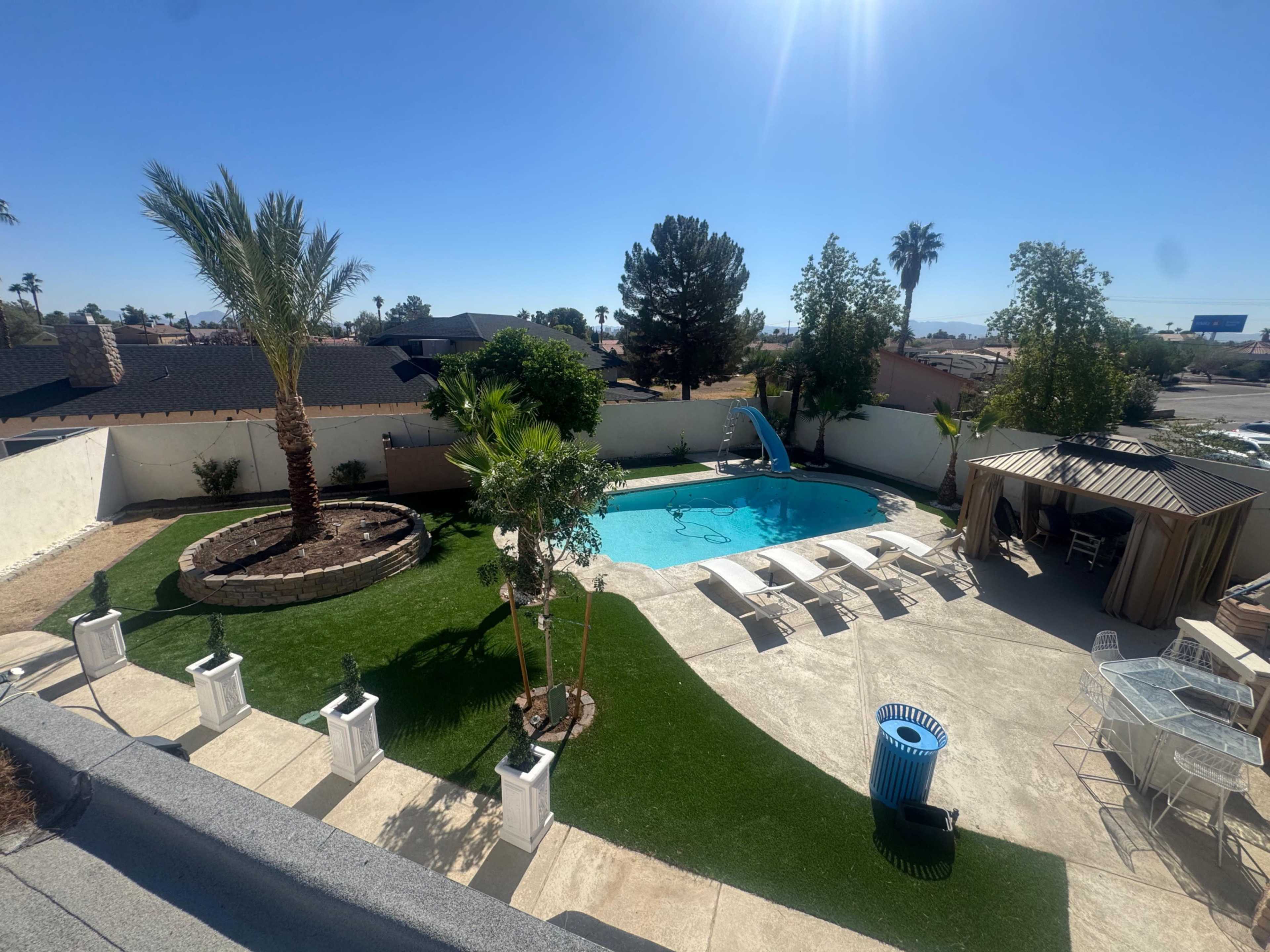 The image shows a backyard with a swimming pool, palm trees, lounge chairs, and a gazebo, surrounded by landscaped grass and a stone wall.