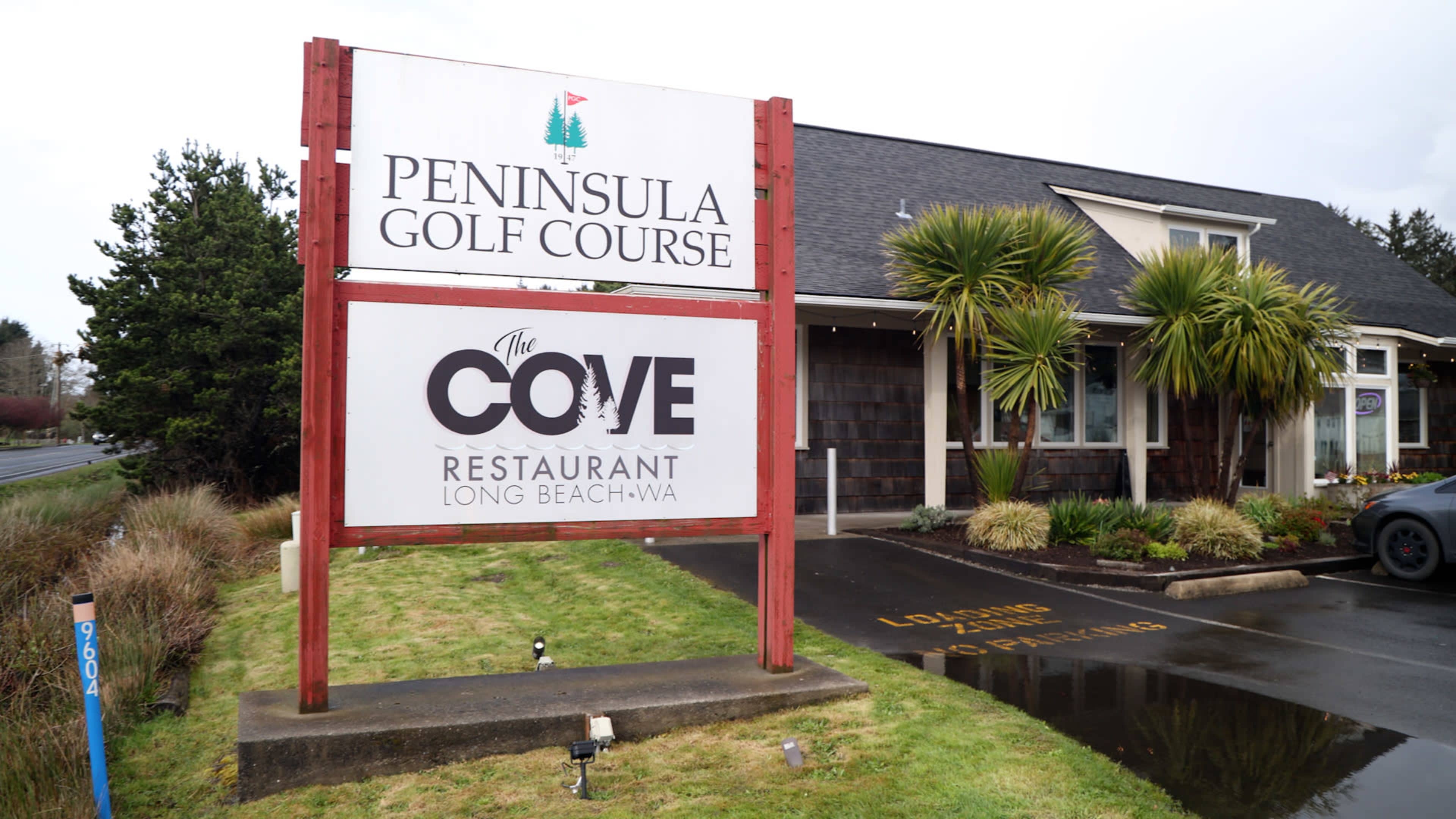The image shows a sign for the Peninsula Golf Course and The Cove Restaurant, located in Long Beach, Washington, in front of a building surrounded by greenery.
